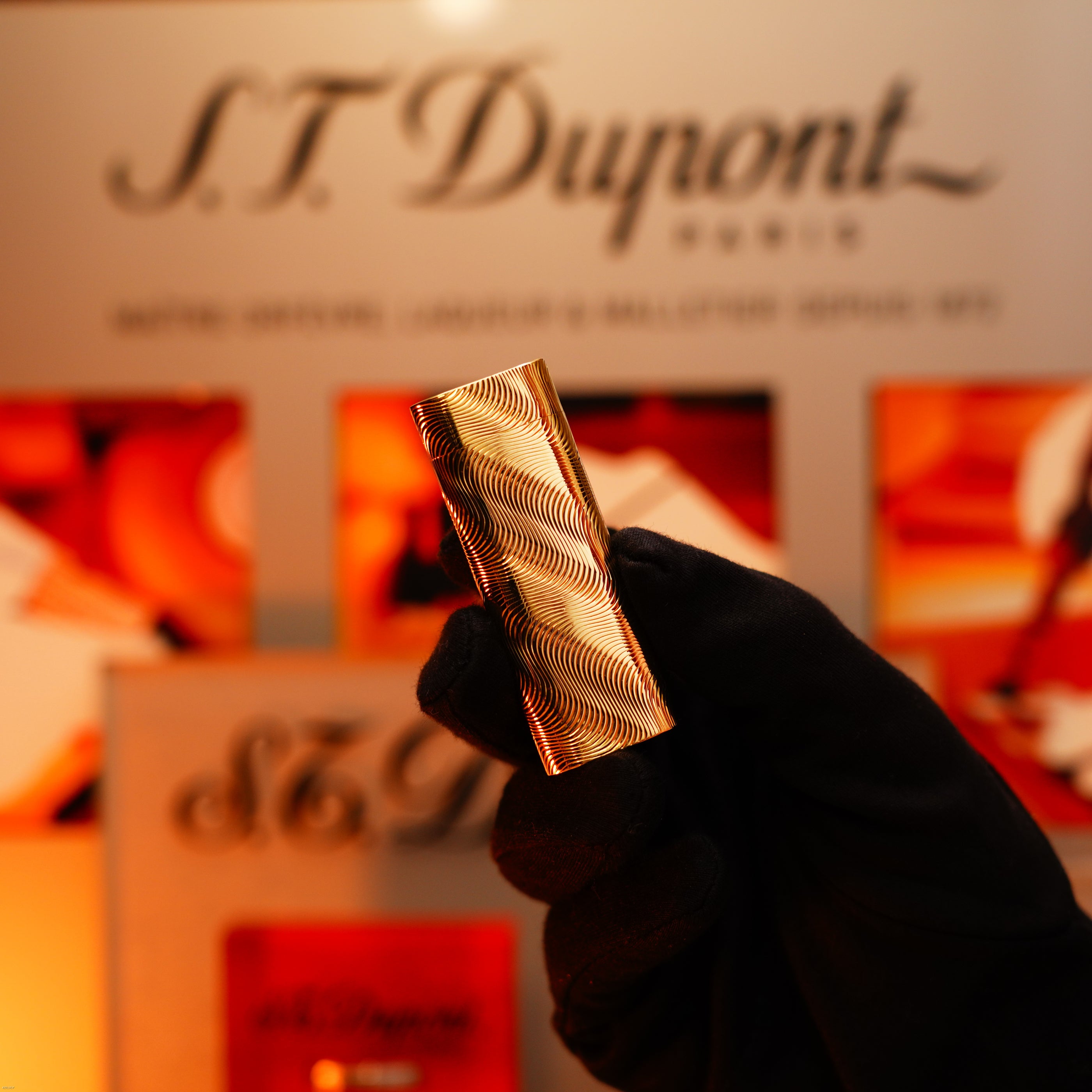 A gloved hand holds a vintage 1970 Lucy Lighter, an extremely rare solid 18K gold lighter with a heavy guilloché pattern and 750 hallmark, in front of a blurred branded display background.