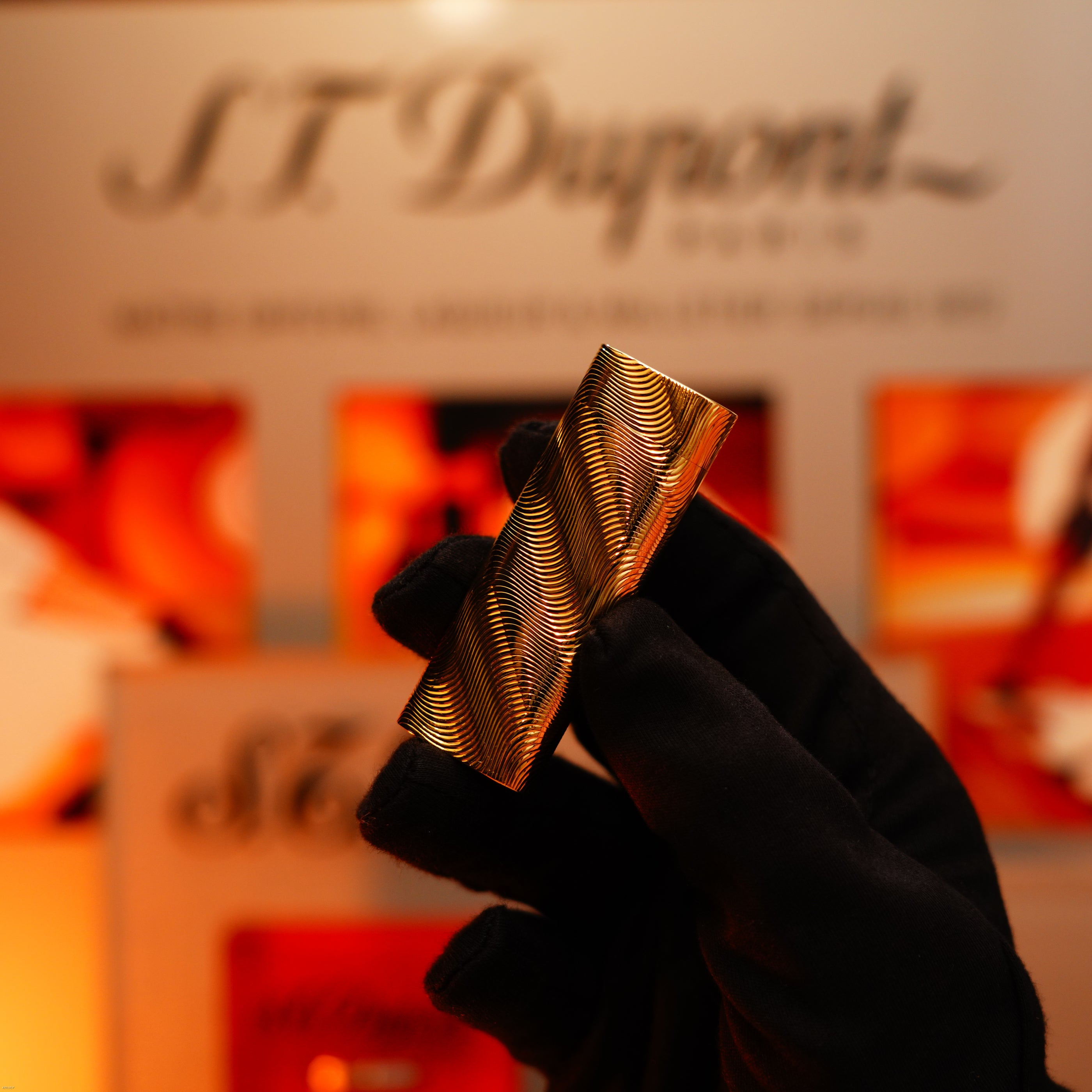A gloved hand holds a Vintage 1970 Cartier Extremely Rare Solid 18K Gold, Heavy Guilloché Pattern, 750 Hallmarked Lucy Lighter in front of a blurred display.