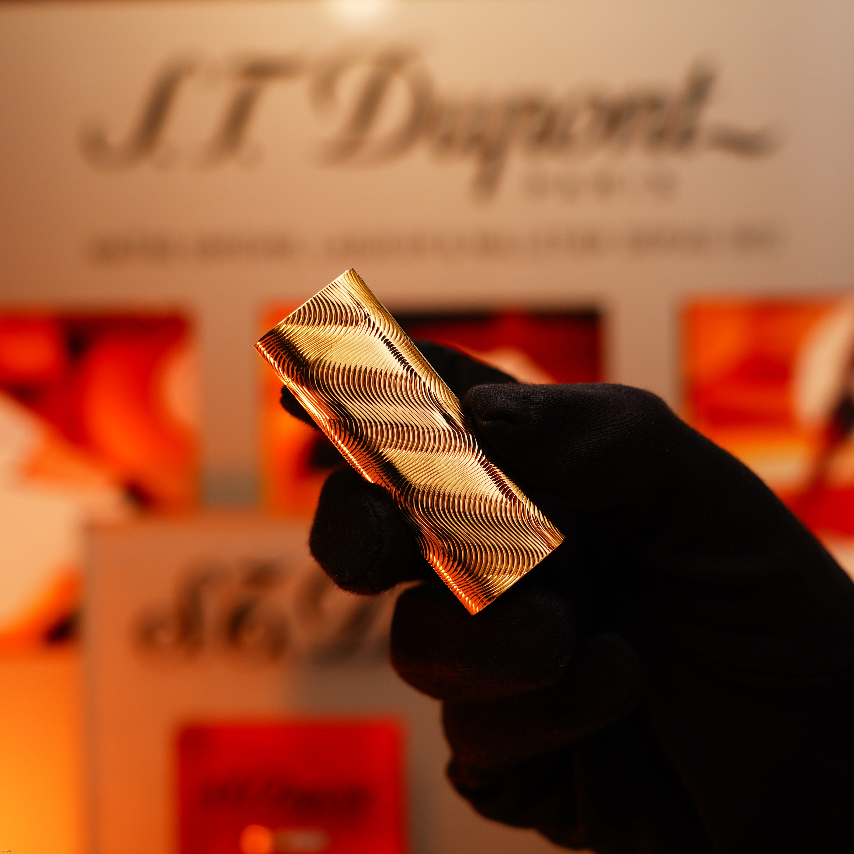 A gloved hand holds a Lucy Lighter Vintage 1970 Cartier Extremely Rare Solid 18K Gold Heavy Guilloché Pattern 750 Hallmarked Lighter, with a blurred branded display in the background.