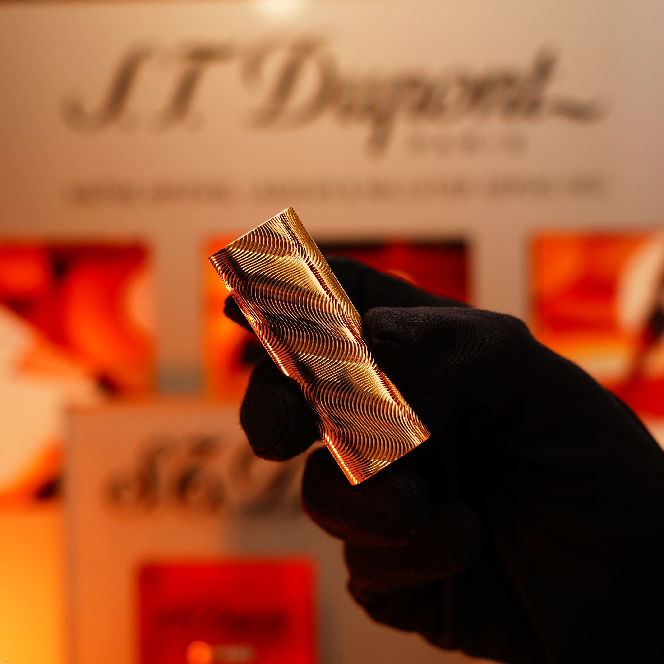 A gloved hand holds a vintage 1970 Cartier Lucy Lighter, an extremely rare solid 18K gold, heavy guilloché pattern (750 hallmarked) lighter, with a blurred display in the background.
