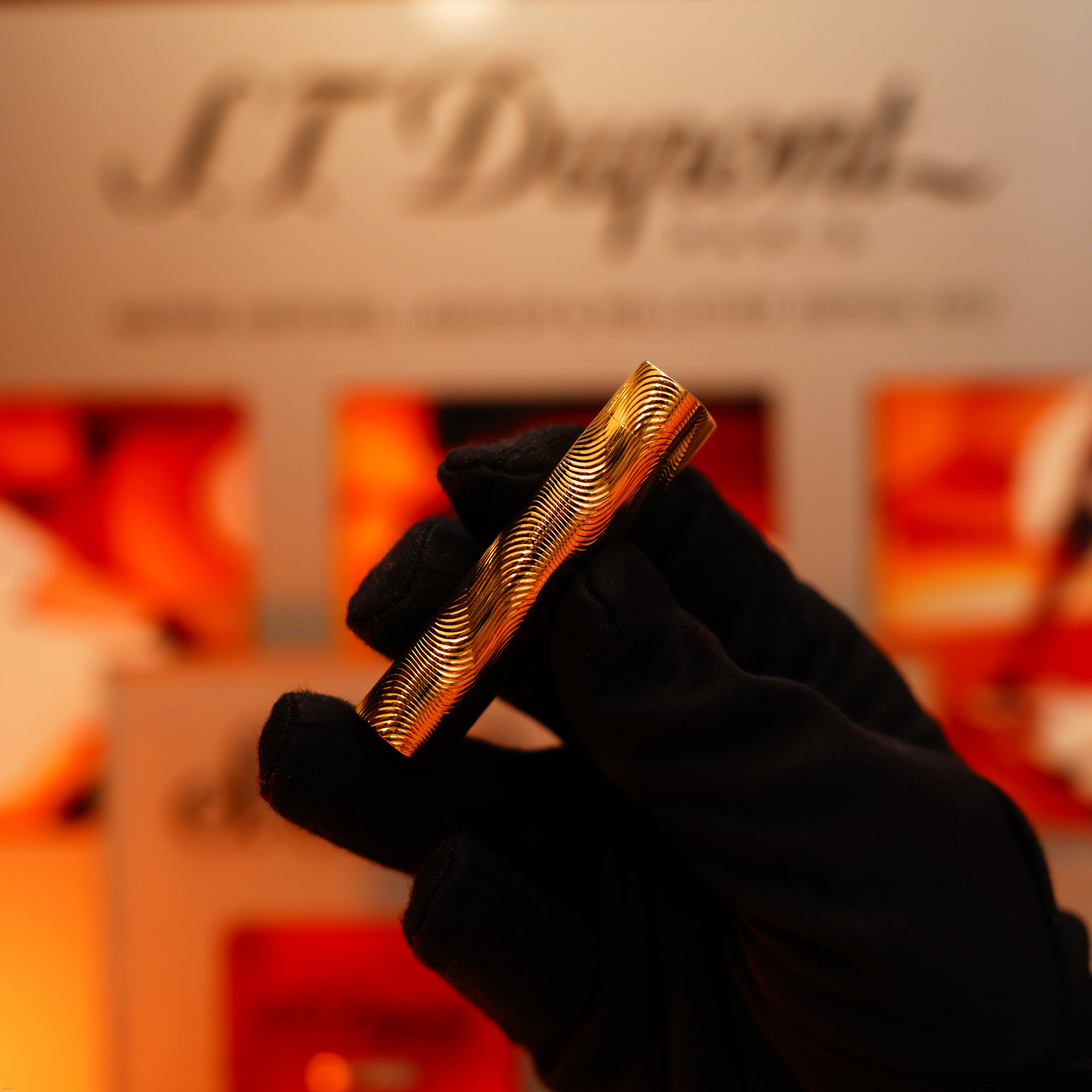 A gloved hand holds a Vintage 1970 Cartier extremely rare solid 18K gold Lucy Lighter with heavy guilloché pattern, 750 hallmarked, in front of a blurred display background.