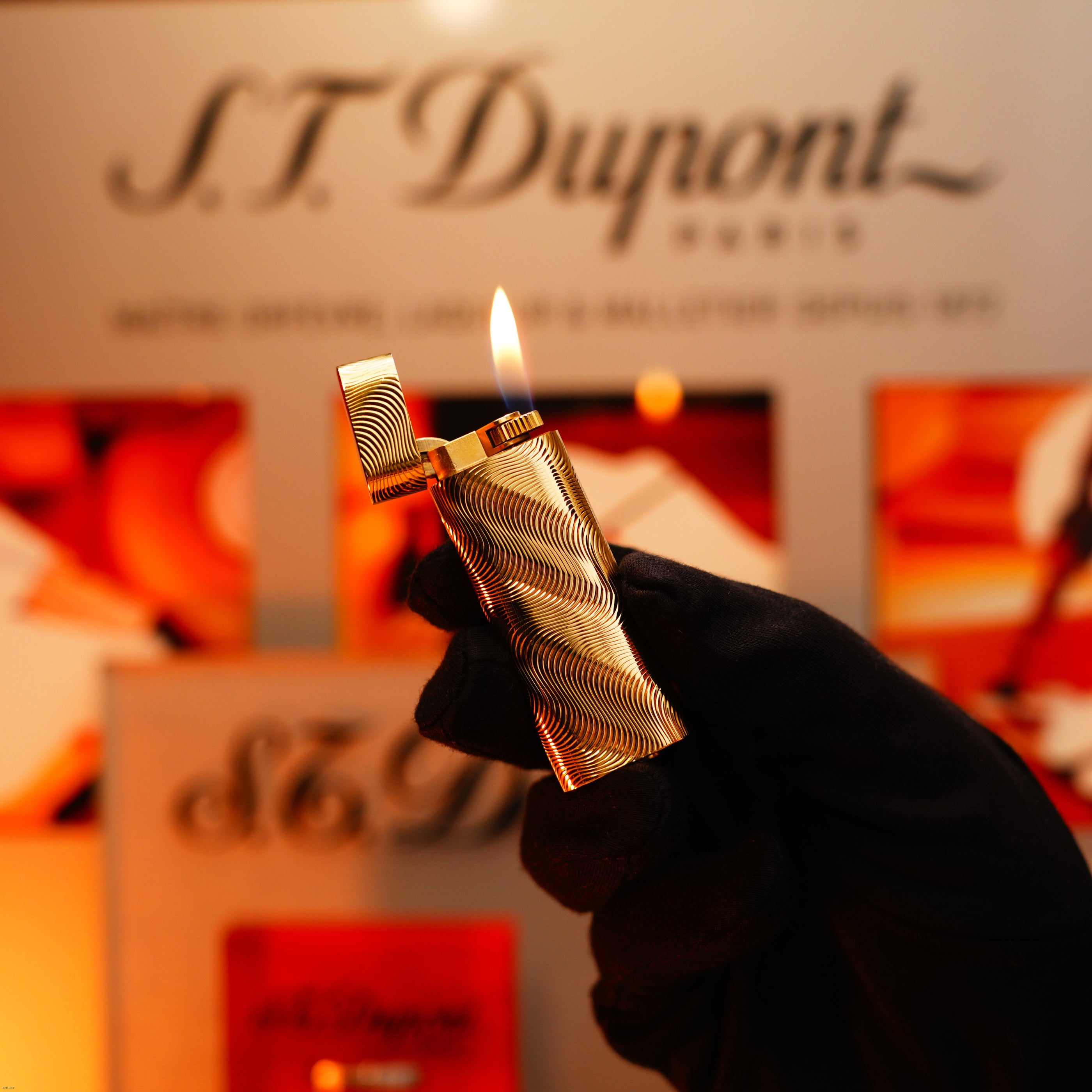 A gloved hand holds a lit Lucy Lighter Vintage 1970 Cartier, an extremely rare solid 18K gold lighter with a heavy guilloché pattern and 750 hallmark, in front of a blurred display.