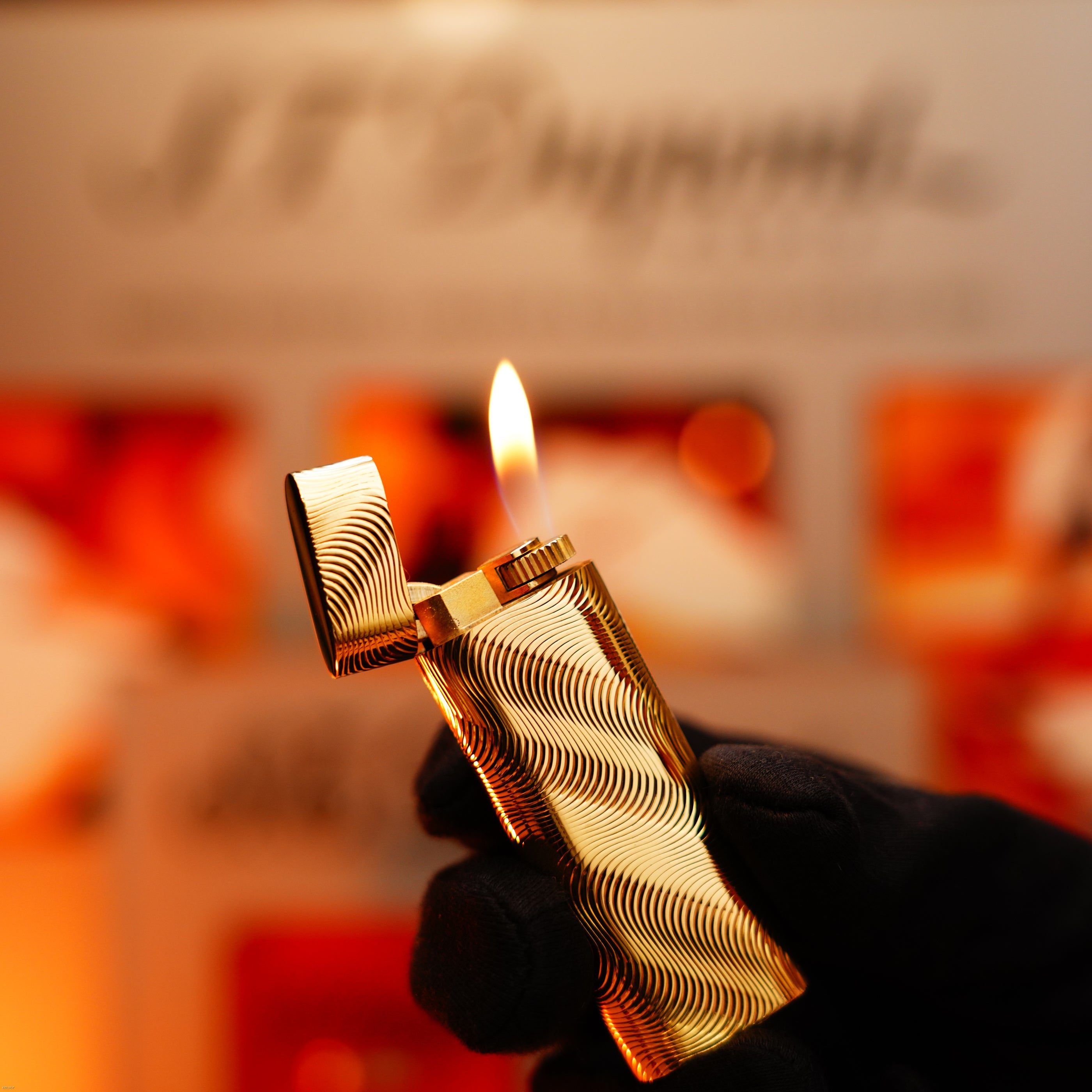 A gloved hand holds a Lucy Lighter Vintage 1970 Cartier Extremely Rare Solid 18K Gold, Heavy Guilloché Pattern, 750 Hallmarked Lighter with its lid open and flame lit, set against a blurred warm-toned background.