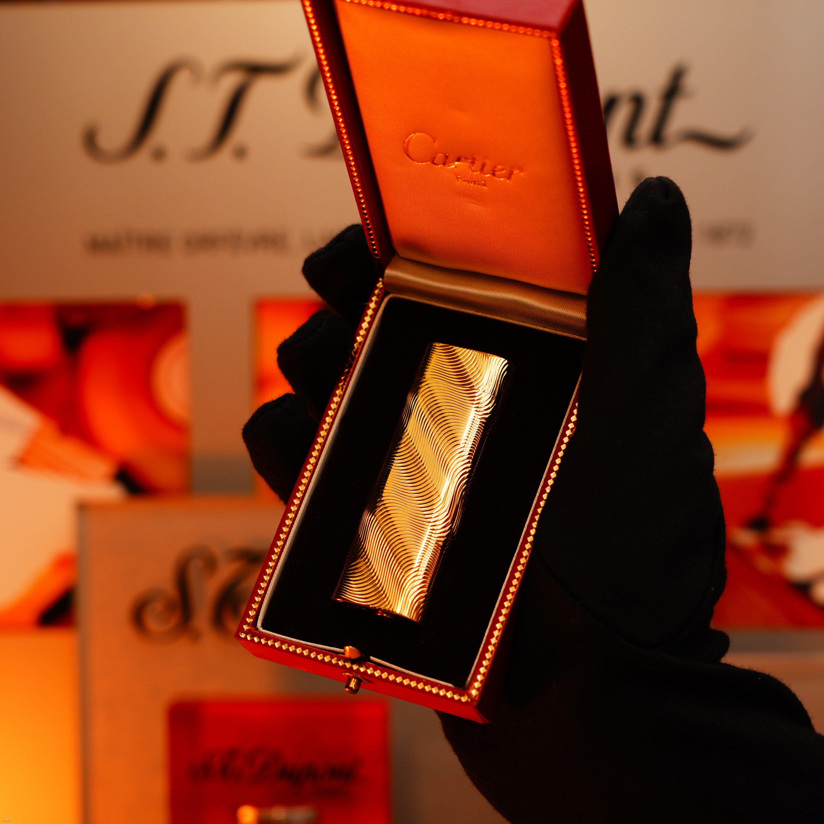 A gloved hand holds an open Lucy Lighter box displaying a Vintage 1970 Cartier Extremely Rare Solid 18K Gold Heavy Guilloché Pattern, 750 Hallmarked Lighter against a blurred background.