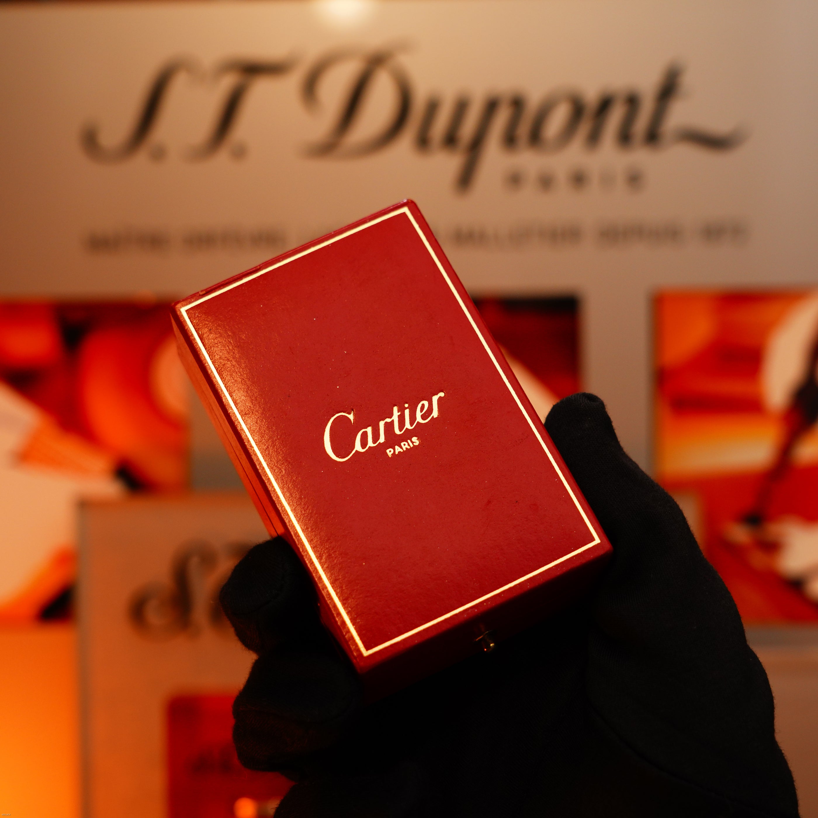 A gloved hand holds a Vintage 1970 Cartier Extremely Rare Solid 18K Gold Guilloché Lighter by Lucy Lighter before a blurred S.T. Dupont Paris display, emphasizing its luxury, rarity, and guaranteed quality.