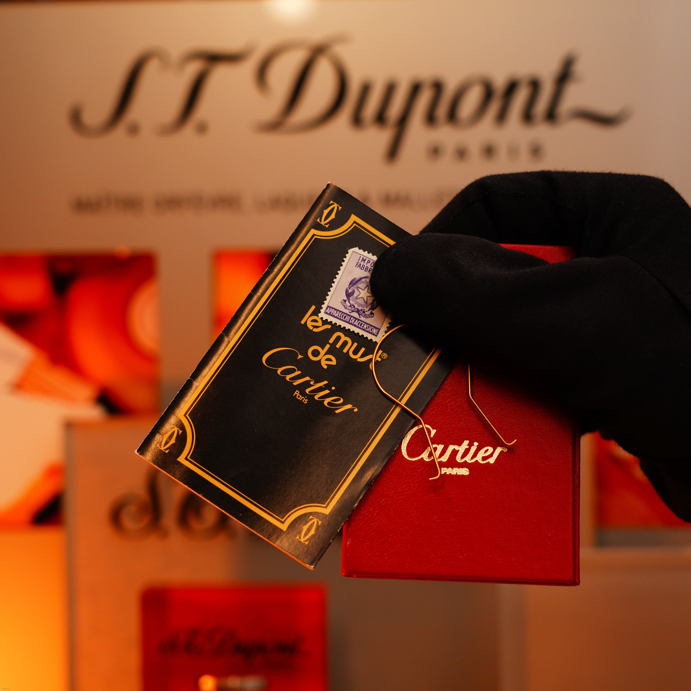 A gloved hand holds a red box and "le must de Cartier" card before an S.T. Dupont Paris display, showcasing the Vintage 1970 Cartier Extremely Rare Solid 18K Gold Heavy Guilloché Pattern 750 Hallmarked Lighter by Lucy Lighter.