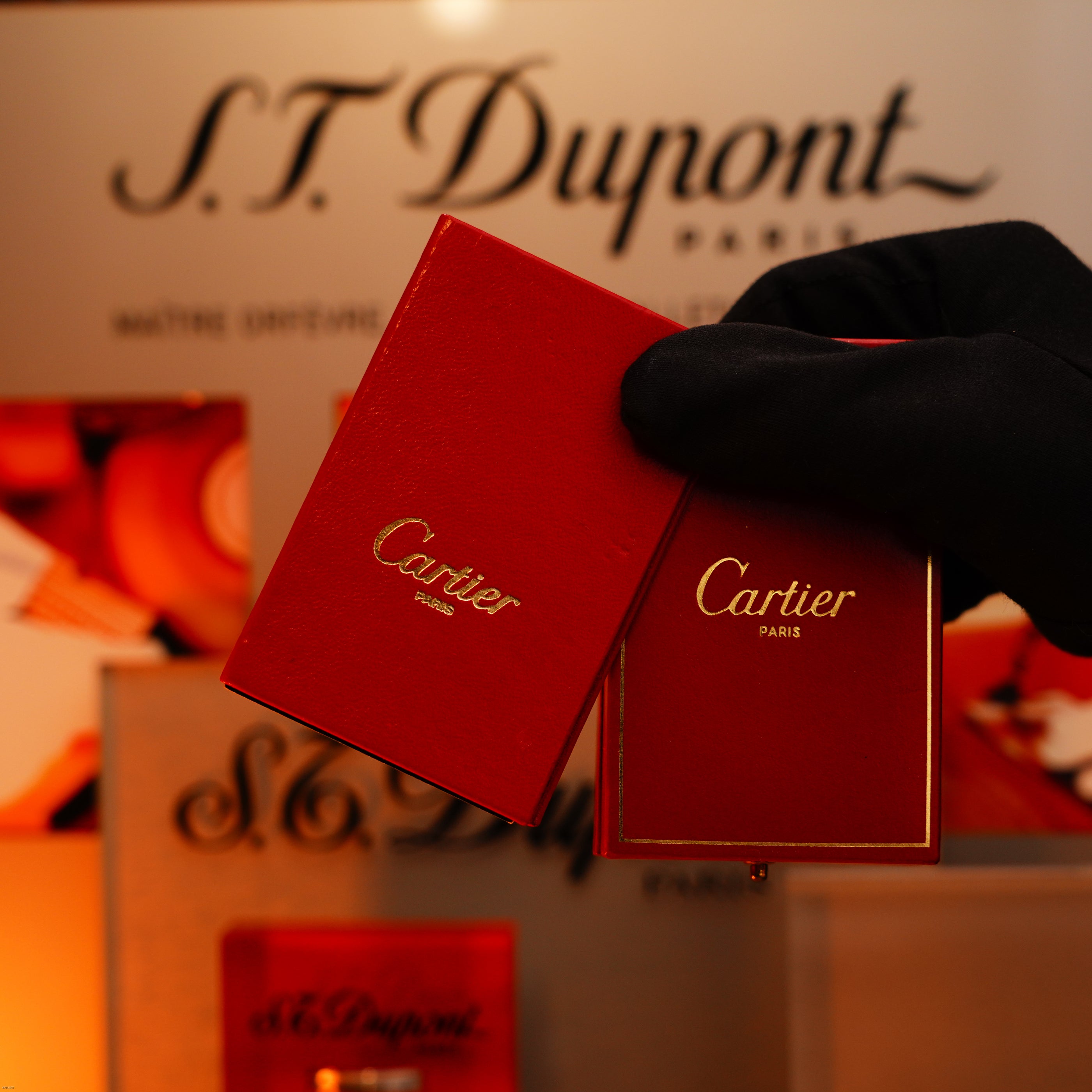 A gloved hand holds two red Cartier boxes before an S.T. Dupont display, evoking the luxury of the Vintage 1970 Cartier Extremely Rare Solid 18K Gold Guilloché Pattern Lighter by Lucy Lighter, 750 hallmarked.