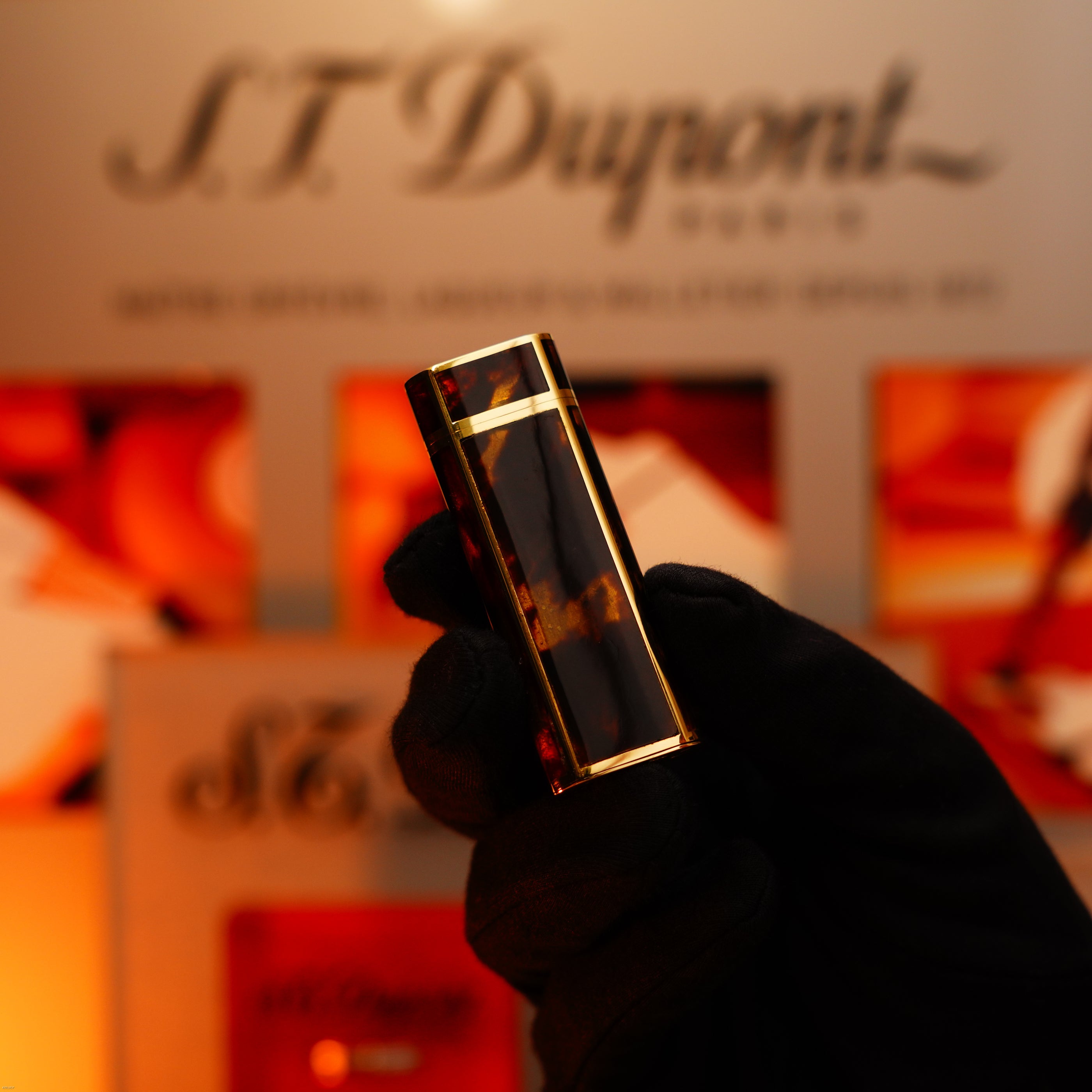 A gloved hand holds the Lucy Lighter Vintage 1970 Limited Cartier Extremely Rare Solid 18K Gold Tiger’s Eye Resin 1/1 Lighter against a blurred background with the Lucy Lighter logo and orange tones.