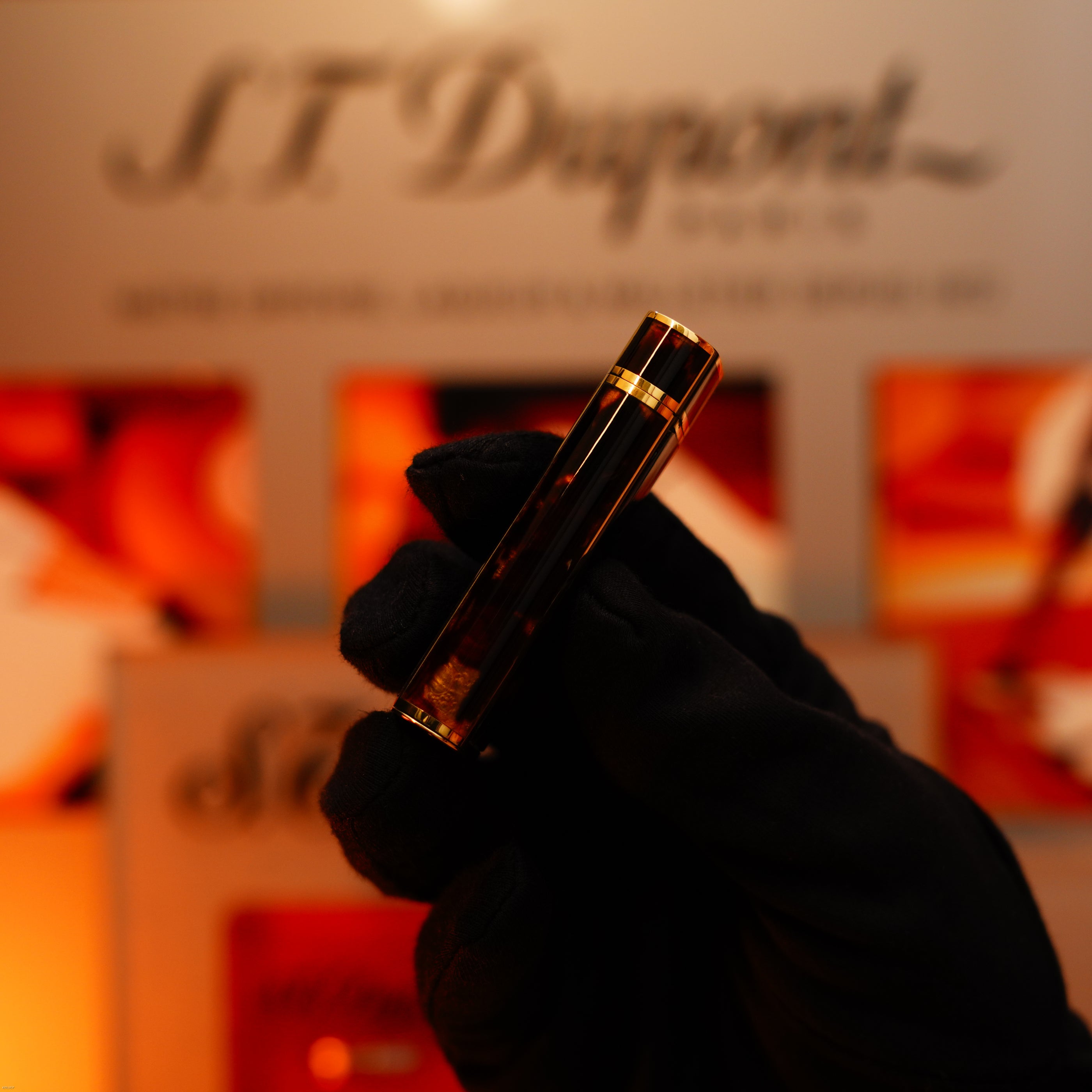 A gloved hand holds the Vintage 1970 Limited Cartier Extremely Rare Solid 18K Gold Tiger’s Eye Resin Lighter by Lucy Lighter, with a blurred display behind, highlighting its unique luxury and expert craftsmanship.