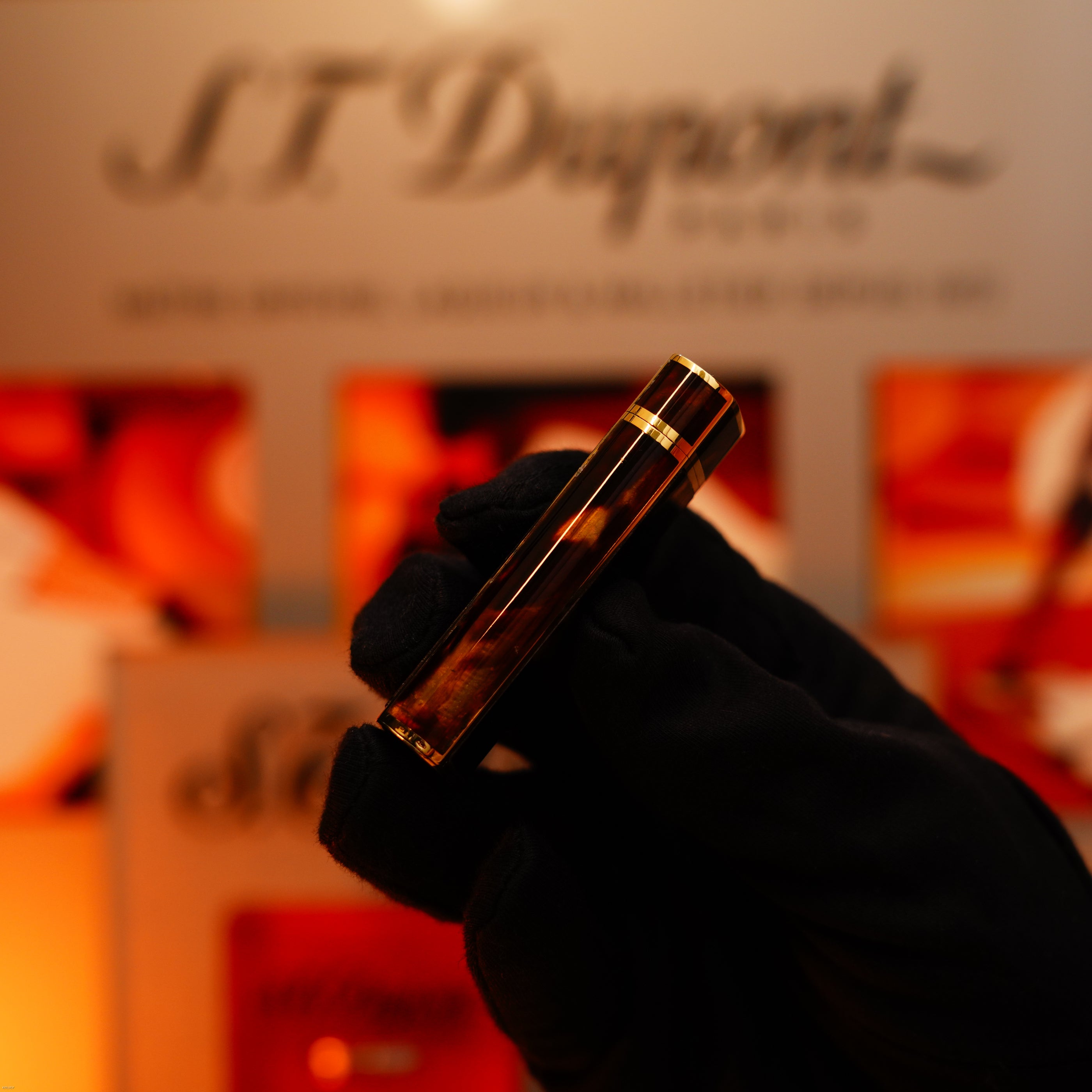 A gloved hand holds a Lucy Lighter Vintage 1970 Limited Cartier solid 18K gold Tiger’s Eye resin lighter (750 hallmarked, 1/1), heavyweight and extremely rare, in front of a blurred display.