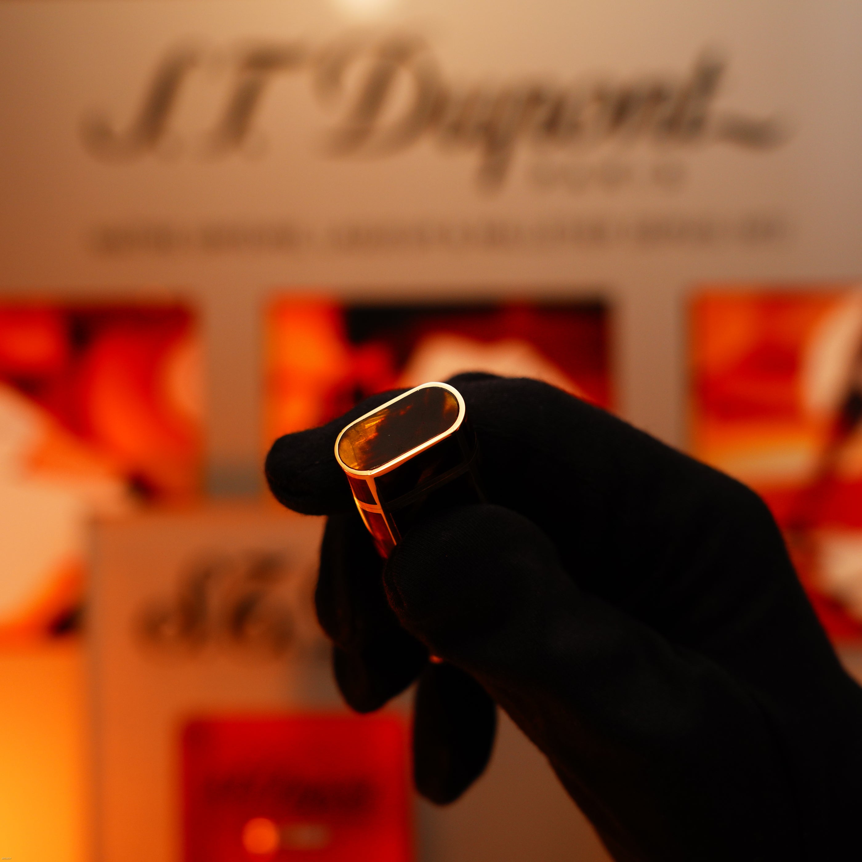 A gloved hand holds a Lucy Lighter Vintage 1970 Limited Cartier solid 18K gold Tiger’s Eye resin lighter, heavyweight and rare, hallmarked 750, in front of a blurred S.T. Dupont display.