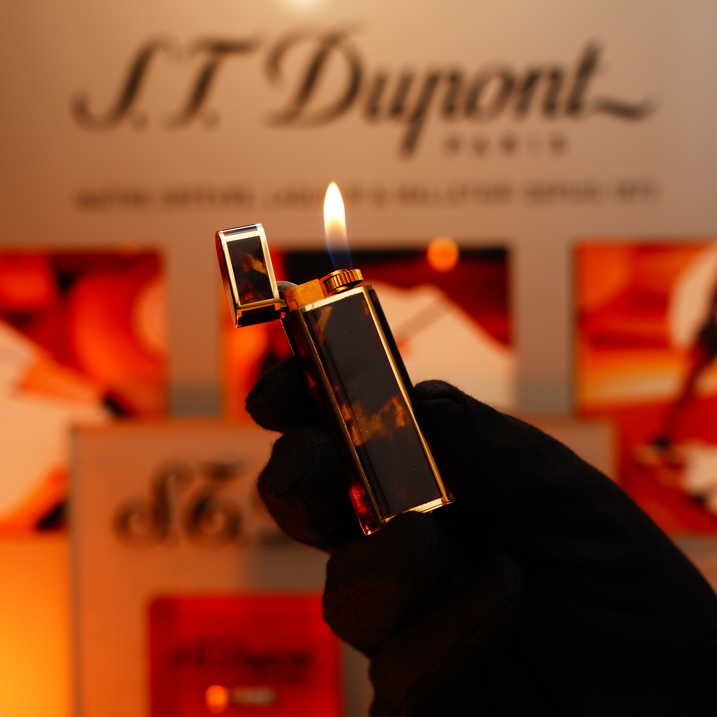 A gloved hand holds a Lucy Lighter Vintage 1970 Limited Cartier Rare Solid 18K Gold Tiger’s Eye Resin 750 Hallmarked 1/1 lighter with the lid open and flame visible, in front of a blurred Lucy Lighter branded background.