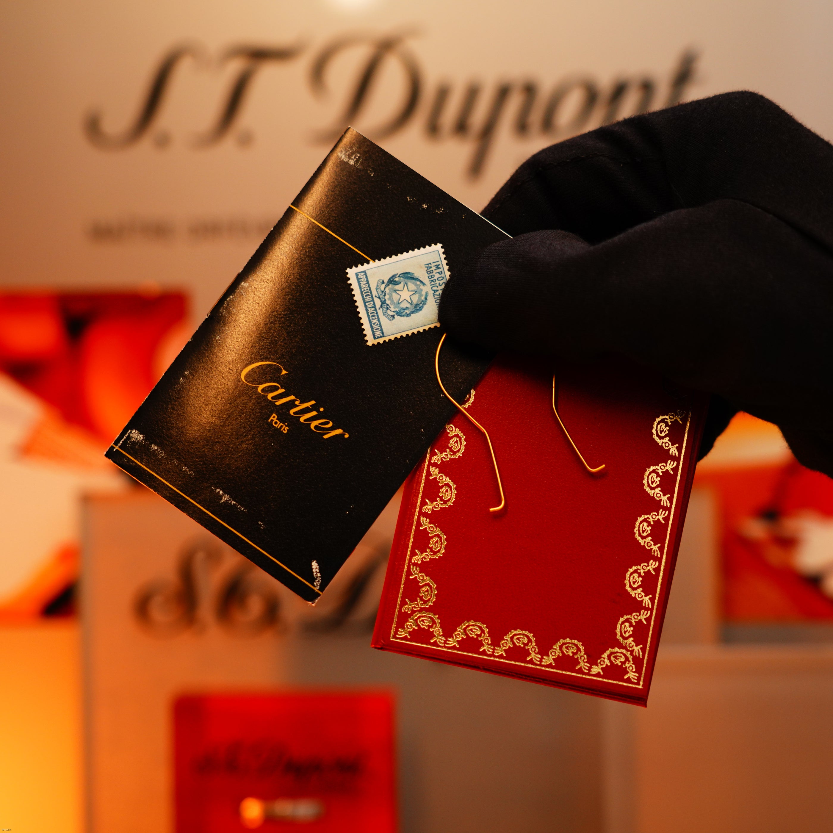 A gloved hand holds chic black and red booklets—classic accessories for your Lucy Lighter Vintage 1970 Limited Cartier 18K Gold Tiger’s Eye Resin 750 Hallmarked Lighter, reflecting style and guaranteed quality.