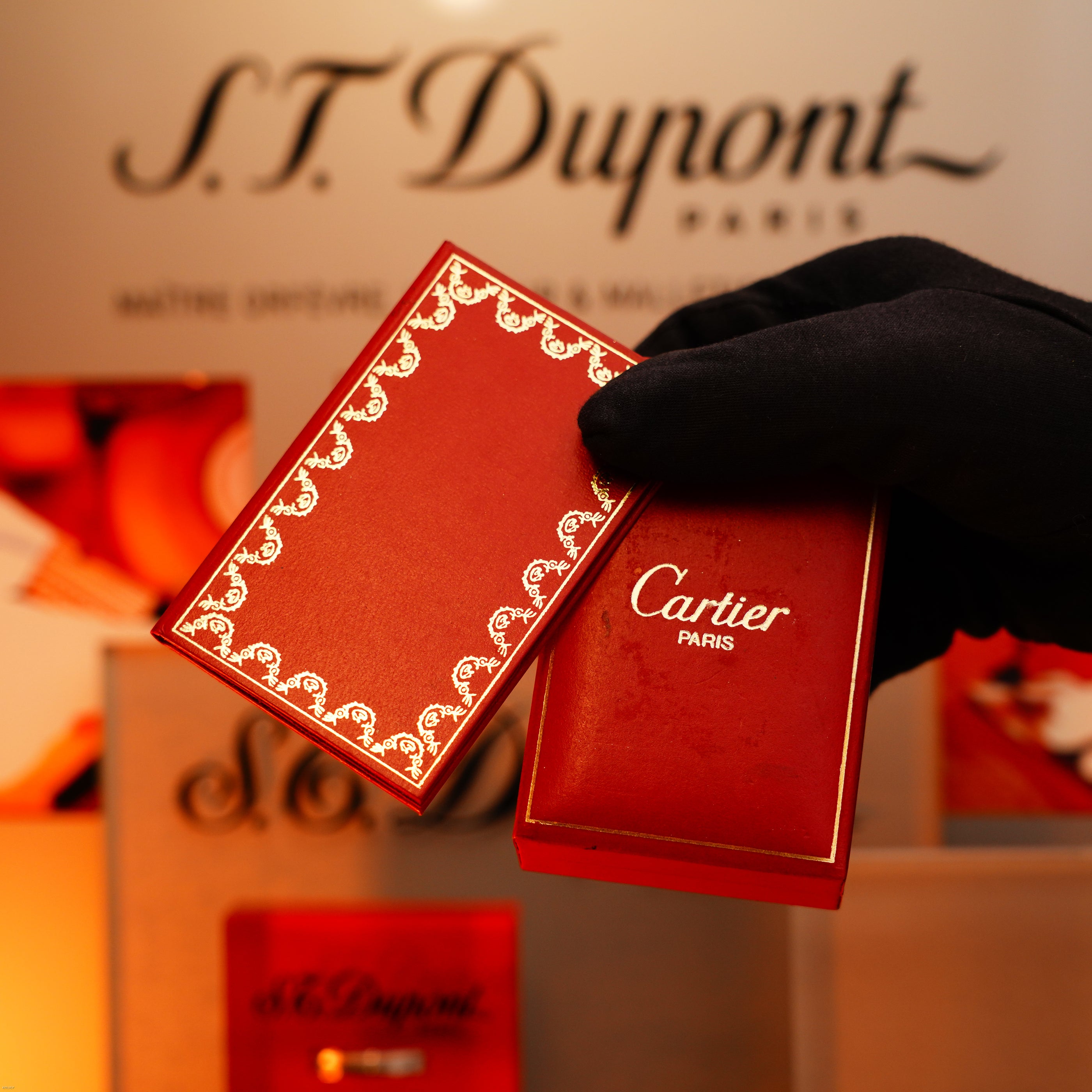 A gloved hand holds two red Lucy Lighter boxes—each possibly containing a Vintage 1970 Limited Cartier 18K Gold Tiger’s Eye Resin Heavyweight Rare Early 1/1 Lighter—set against branded luxury packaging for a mark of exclusivity and quality.