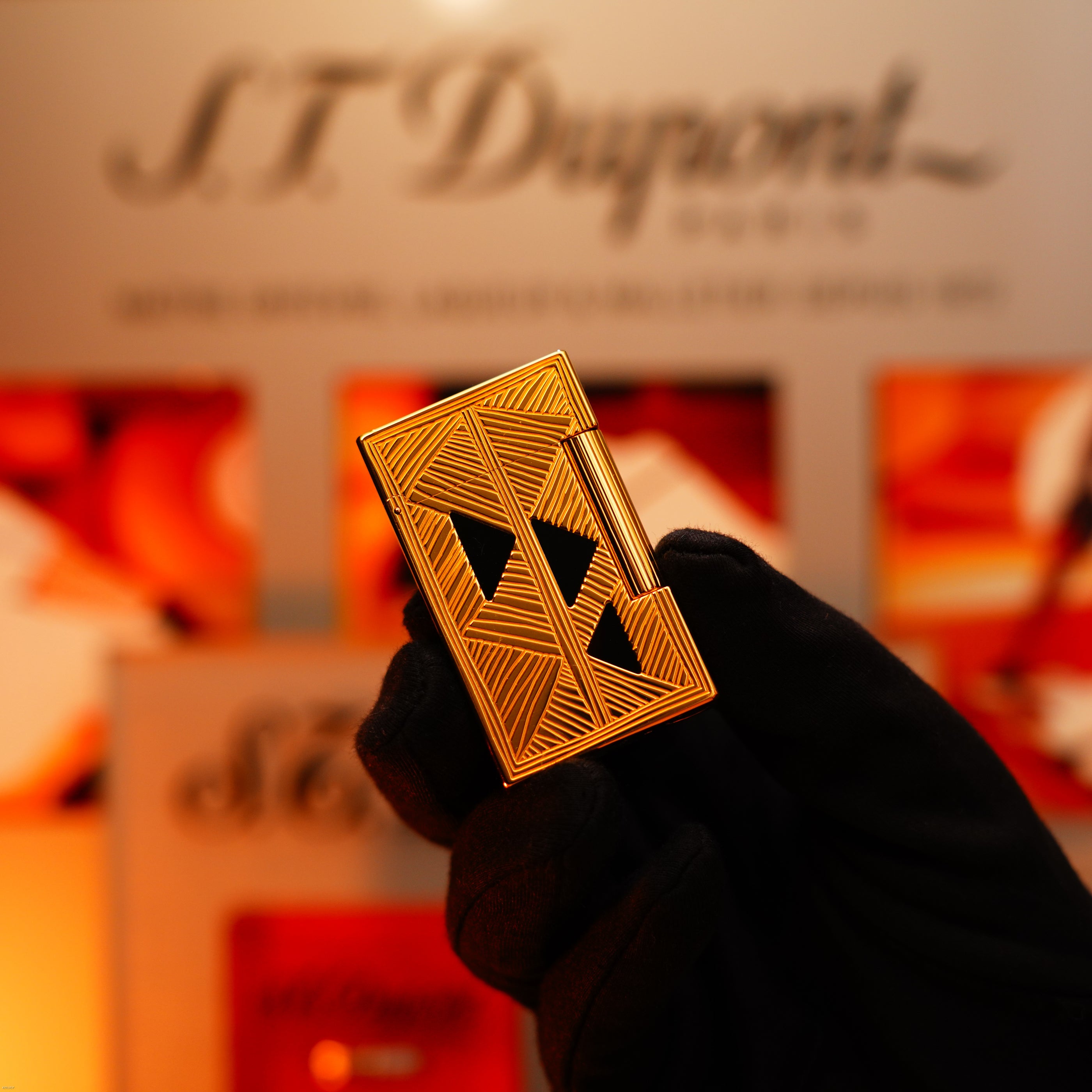 A gloved hand holds a Vintage 2000 St Dupont Limited Edition Heavy 24k Gold Finish Africa Natural Lacquer Large Ligne 2 Lighter by Lucy Lighter, set against a blurred branded background.