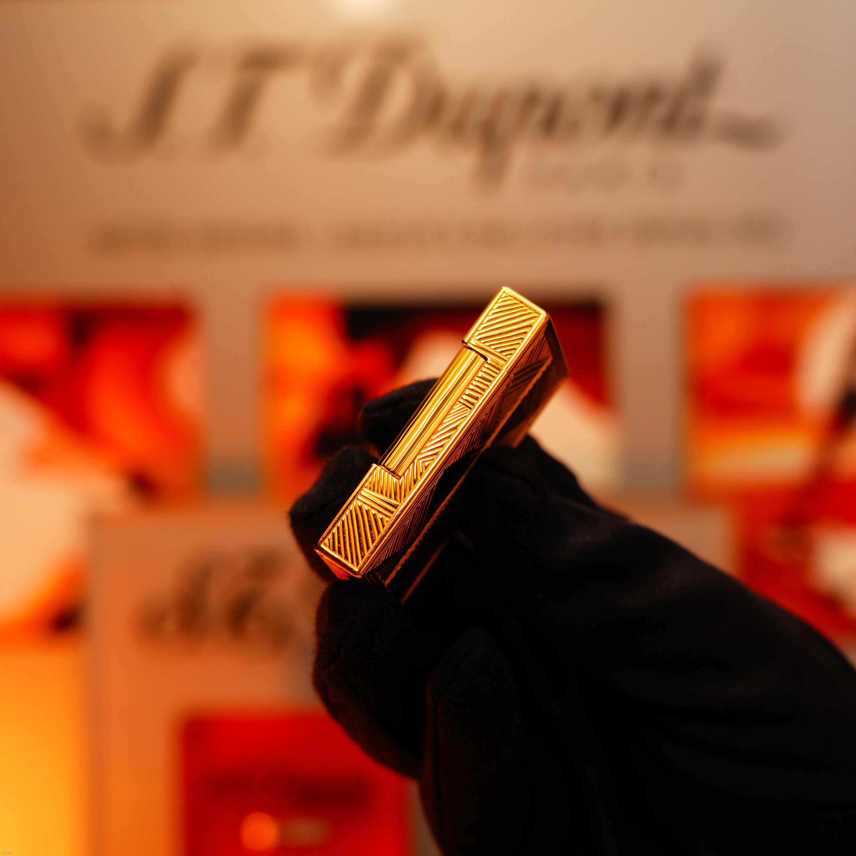 A gloved hand holds a Lucy Lighter Vintage 2000 St Dupont Limited Edition Large Ligne 2 Lighter with heavy 24k gold finish and Africa natural lacquer; a blurred S.T. Dupont sign and display are visible in the background.