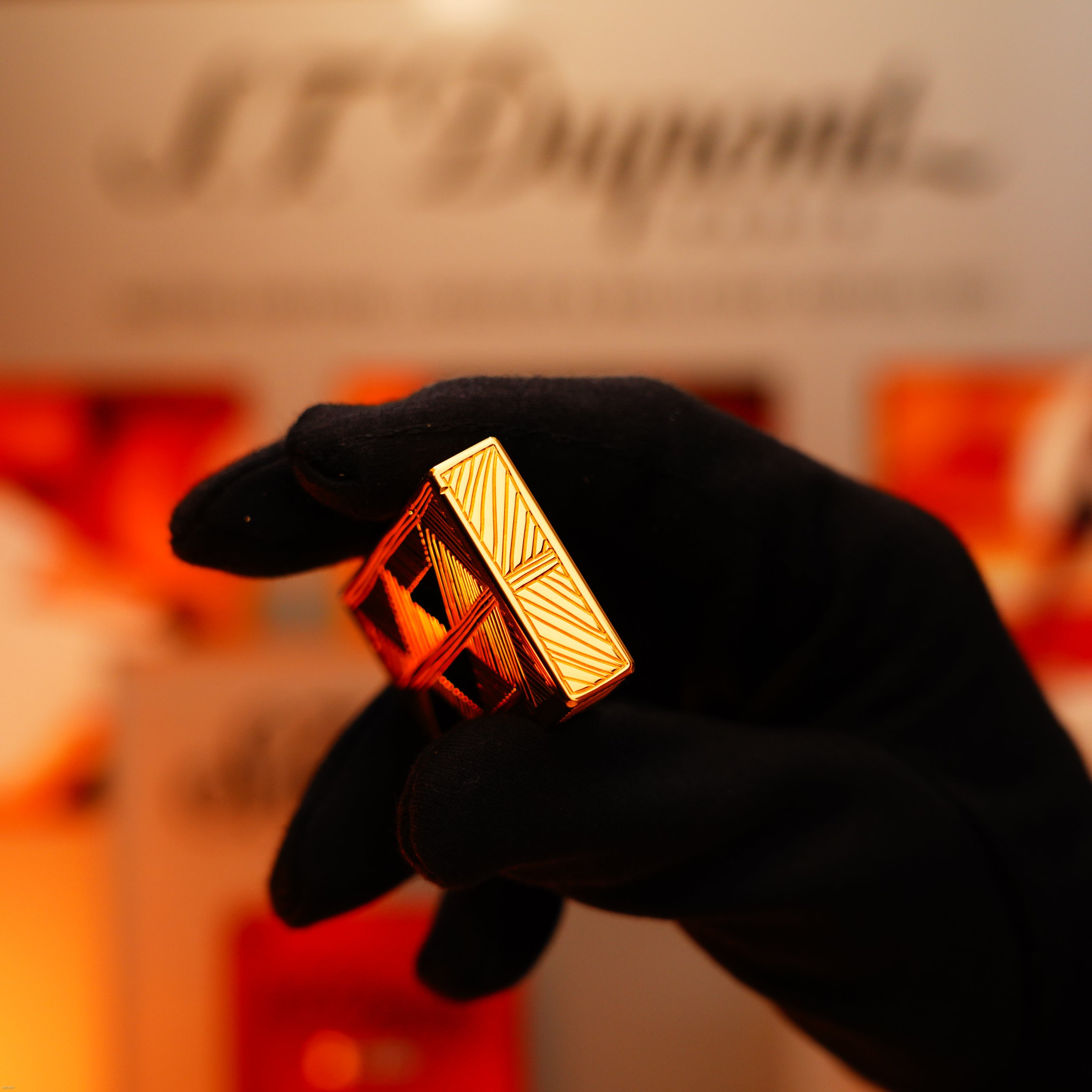 A gloved hand holds a Lucy Lighter Vintage 2000 Limited Edition Heavy 24k Gold Finish Africa Natural Lacquer Large Ligne 2 lighter, with its luxurious geometric design and a blurred "S.T. Dupont Paris" sign in the background.