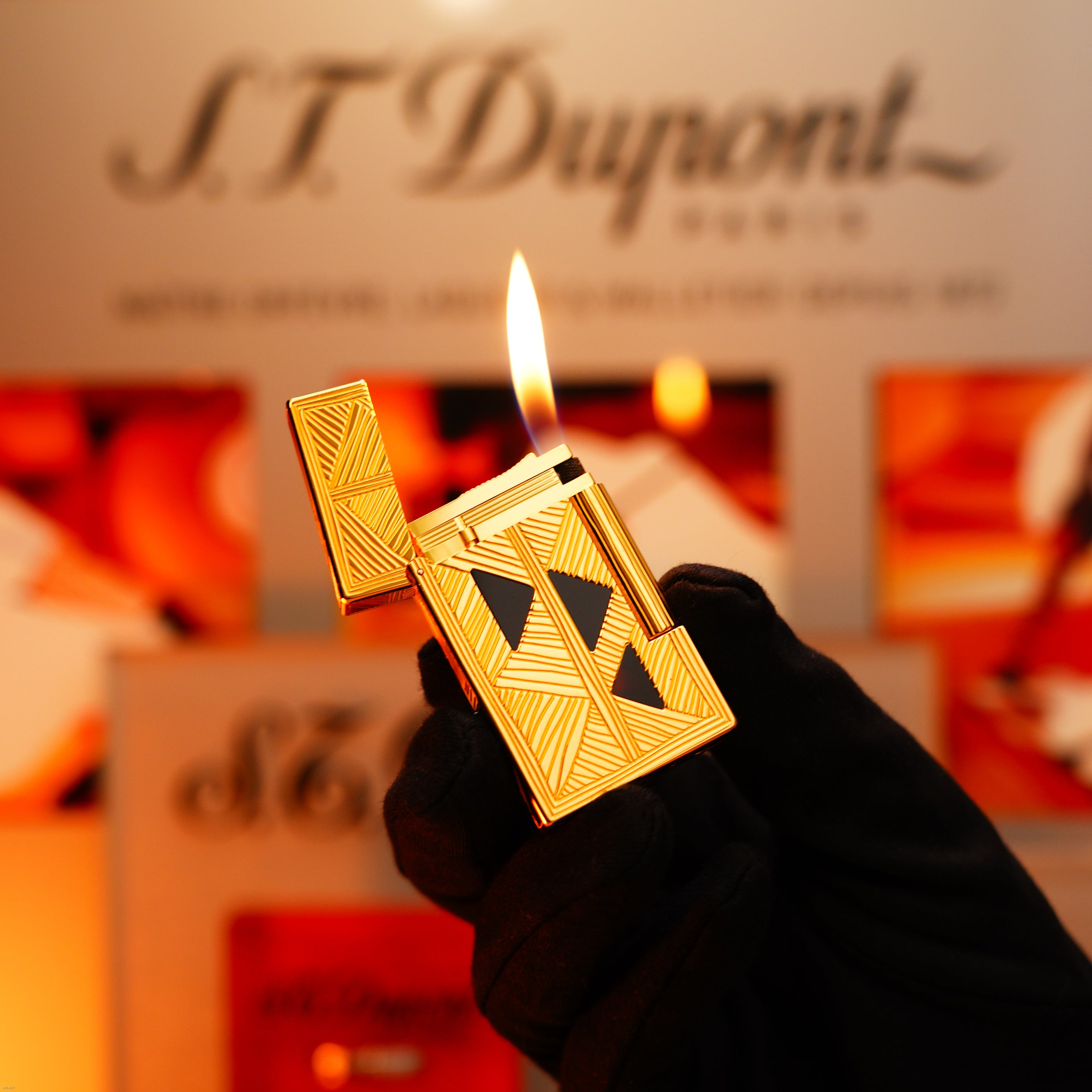 A gloved hand holds a lit Lucy Lighter Vintage 2000 Limited Edition Large Ligne 2 lighter in heavy 24k gold finish with Africa natural lacquer; its branded packaging appears blurred in the background.
