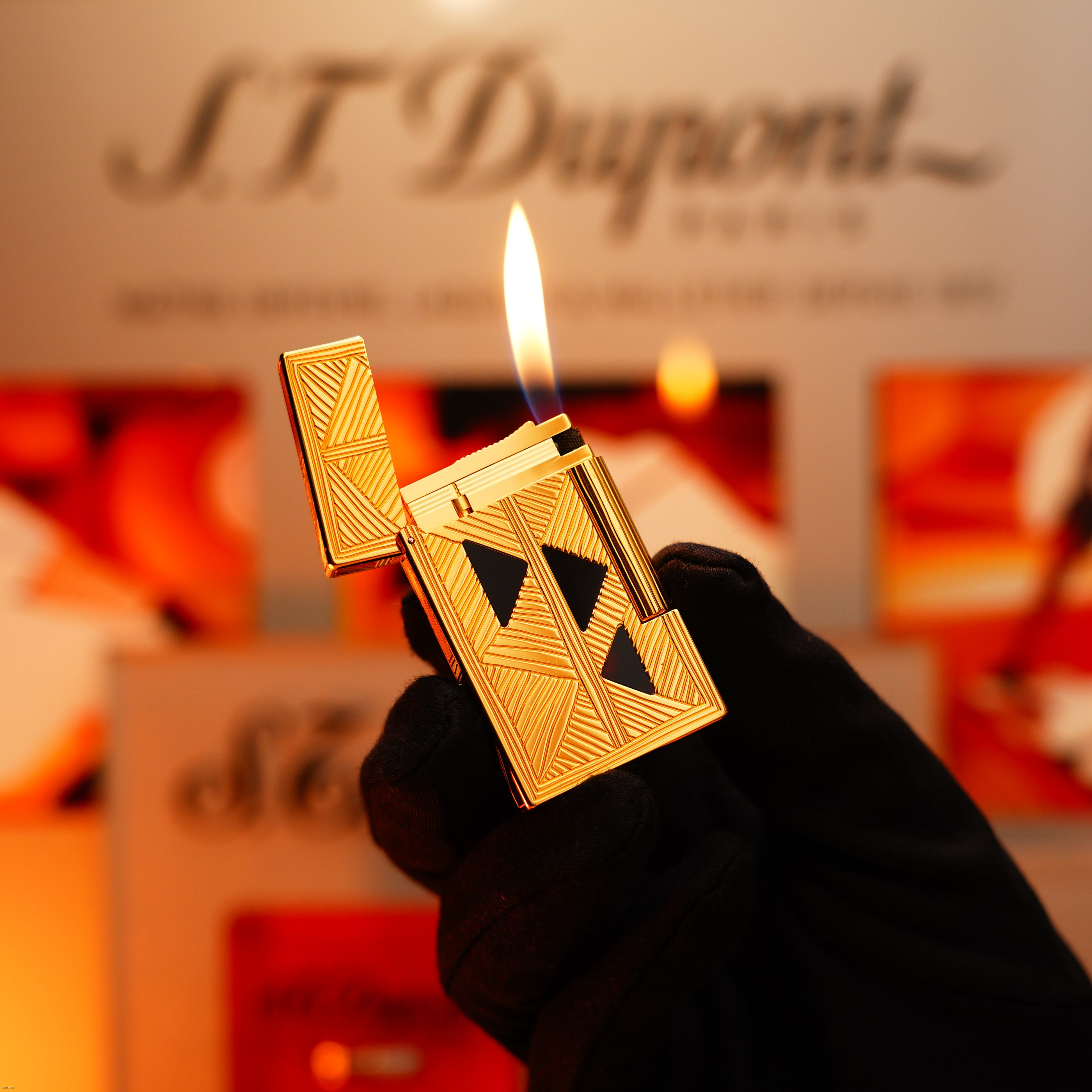 A gloved hand holds a Lucy Lighter Vintage 2000 St Dupont Limited Edition Heavy 24k Gold Finish Africa Natural Lacquer Large Ligne 2 lighter, open with a visible flame against a blurred branded background.