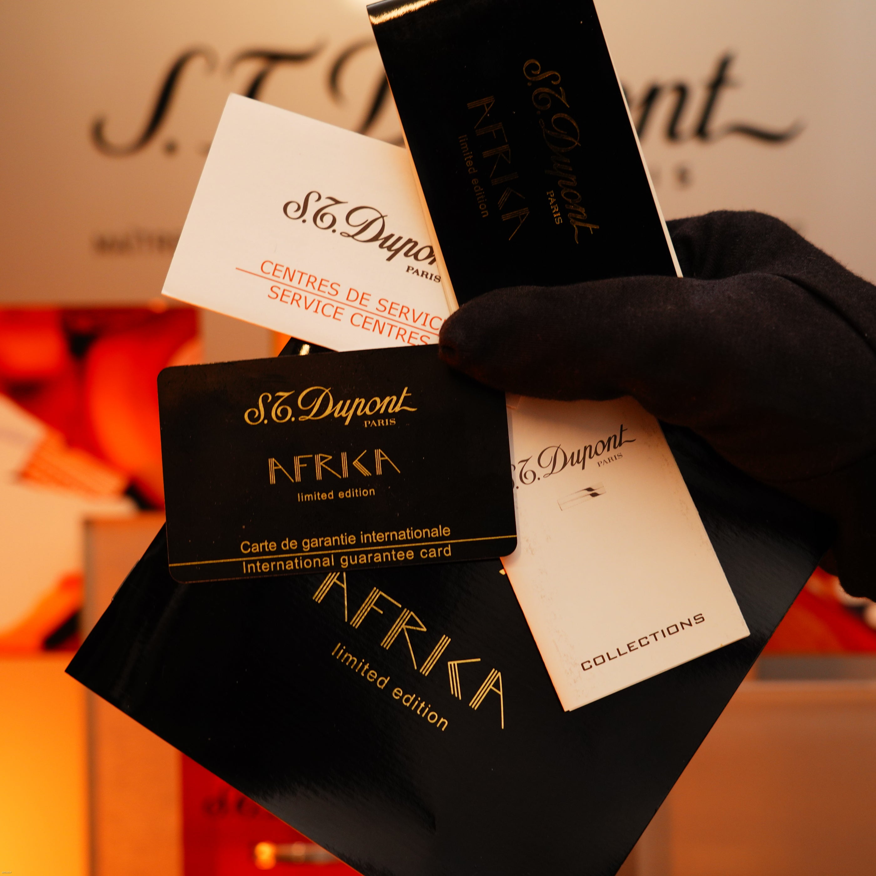 A gloved hand holds several Lucy Lighter cards and booklets, including a warranty card for the Vintage 2000 Limited Edition Heavy 24k Gold Finish Africa Natural Lacquer Large Ligne 2 Lighter, against a blurred display background.