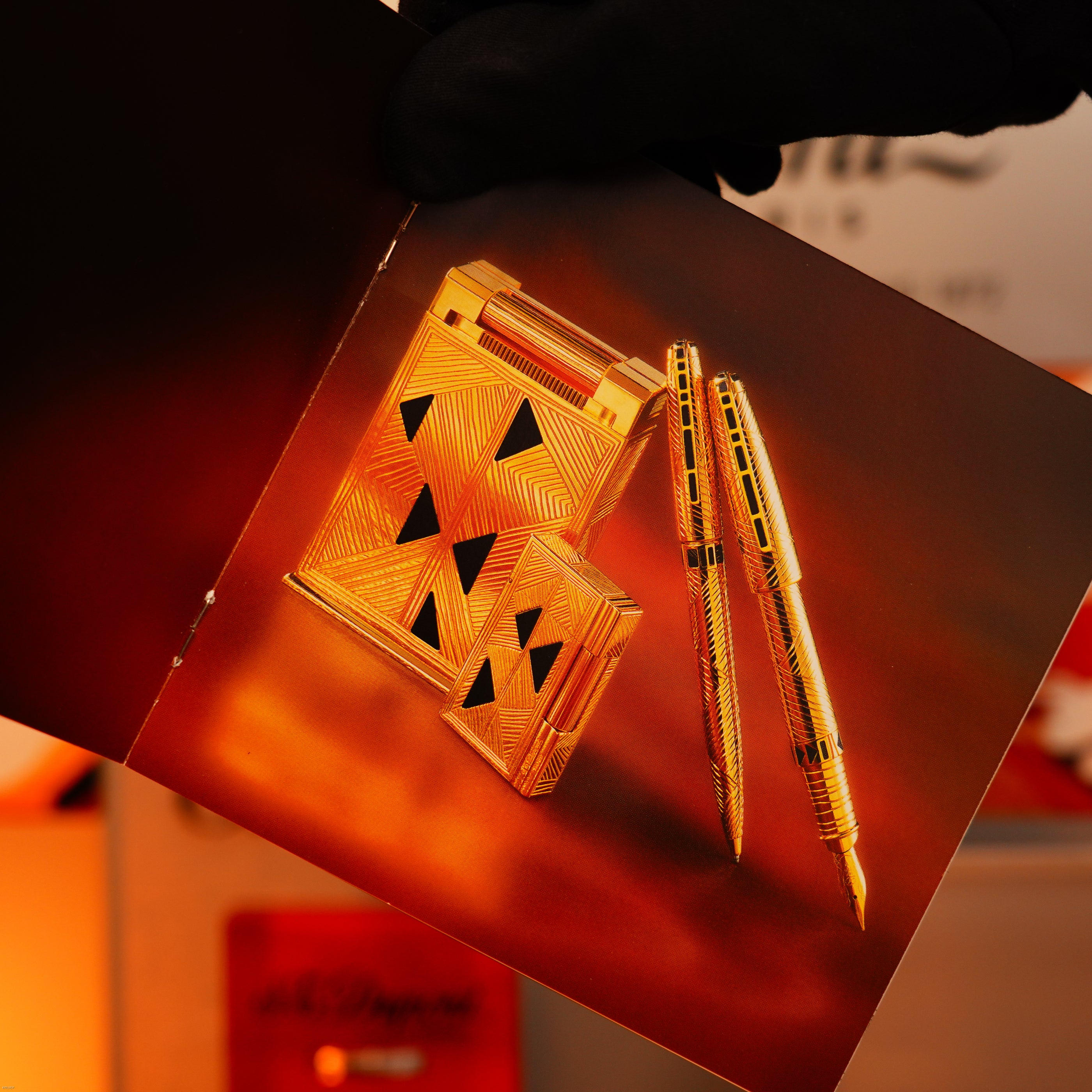 A gloved hand holds a catalog page featuring Lucy Lighter's Vintage 2000 St Dupont Limited Edition Heavy 24k Gold Finish Africa Natural Lacquer Large Ligne 2 Lighter and gold-colored luxury pens on an amber background.