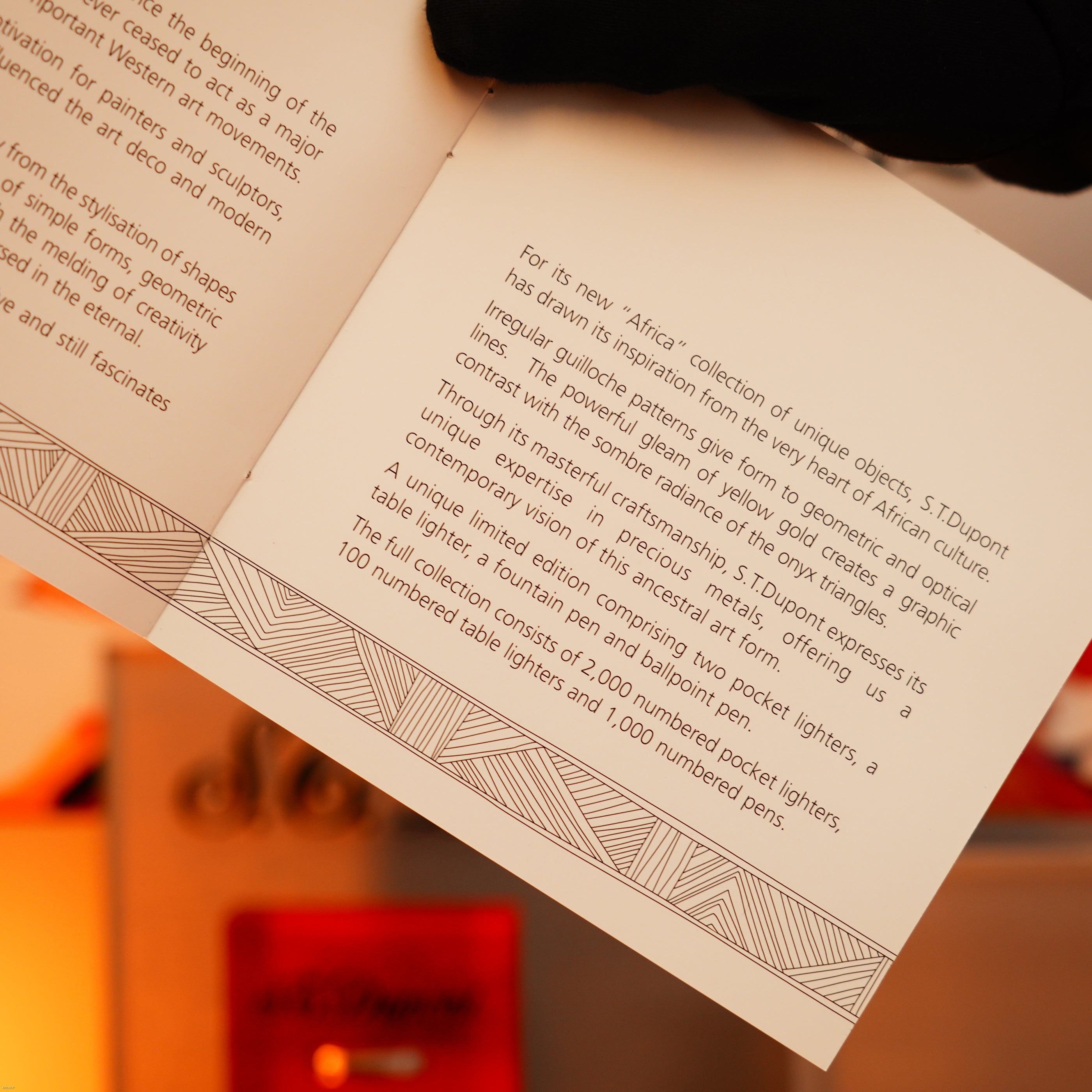 A close-up of a booklet page details the Lucy Lighter Vintage 2000 St Dupont Limited Edition Heavy 24k Gold Finish Africa Natural Lacquer Large Ligne 2 Lighter, with geometric borders and blurred red-gold accents—showcasing vintage luxury.