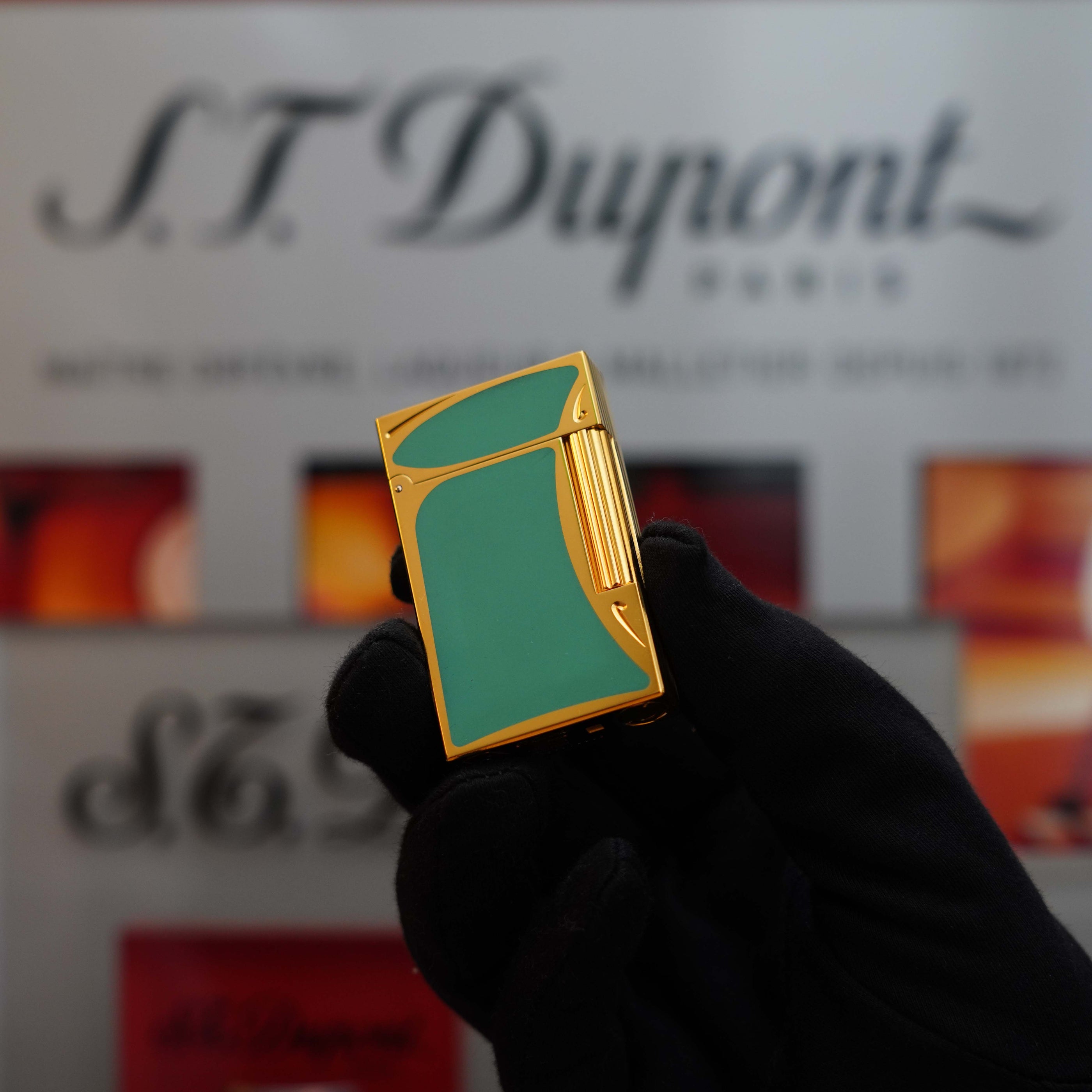 A gloved hand holds a gold and green S.T. Dupont Vintage 1993 rare Art Nouveau early Limited Editions 24k Gold finish natural Green lacquer ligne 2 lighter with a blurred S.T. Dupont logo and text in the background, showcasing its exquisite 24k gold finish.