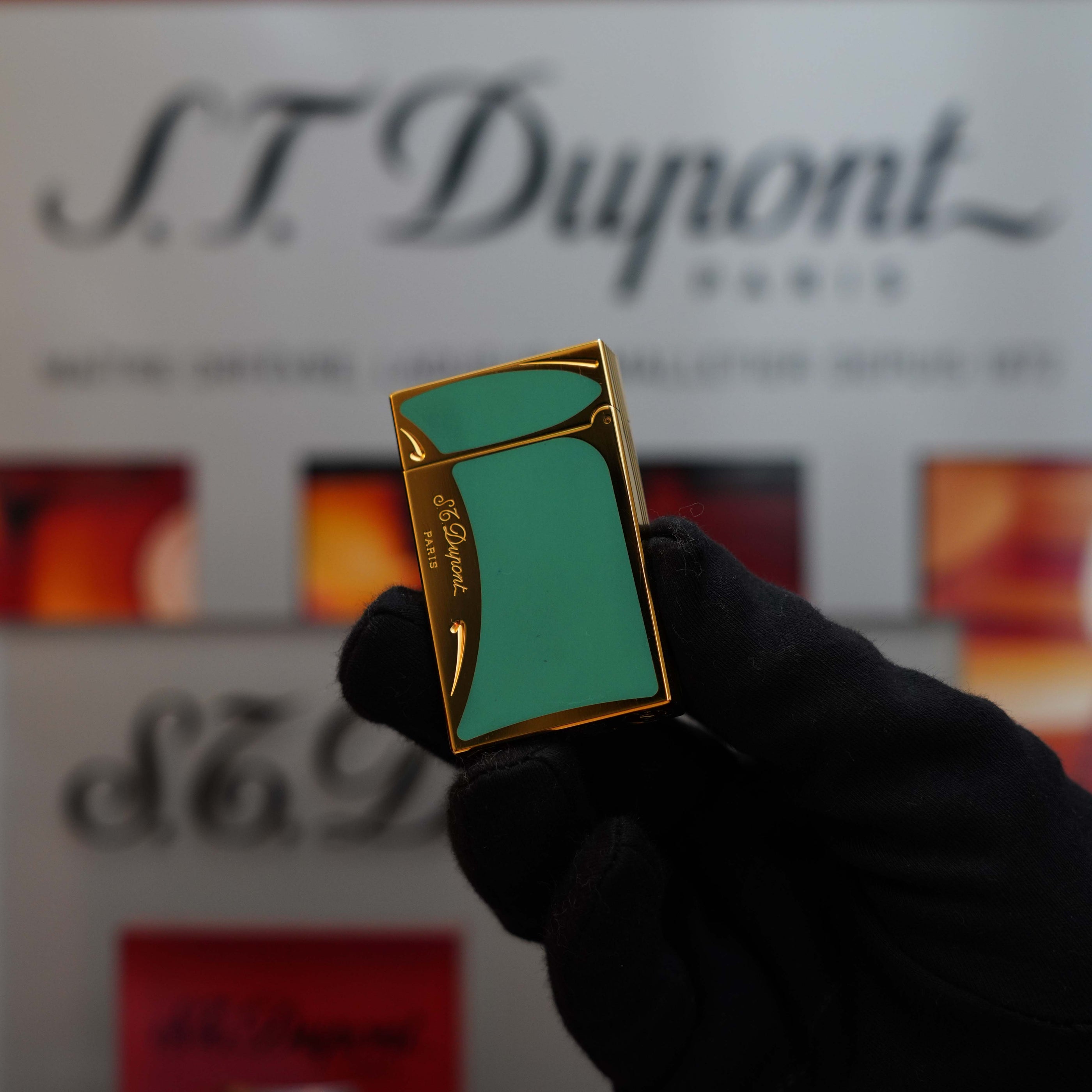 A gloved hand holds a turquoise and gold Vintage 1993 rare Art Nouveau early Limited Editions 24k Gold finish natural Green lacquer ligne 2 lighter. An S.T. Dupont logo and various images of vintage lighters are blurred in the background, showcasing St Dupont Limited Editions with exquisite 24k gold finish.