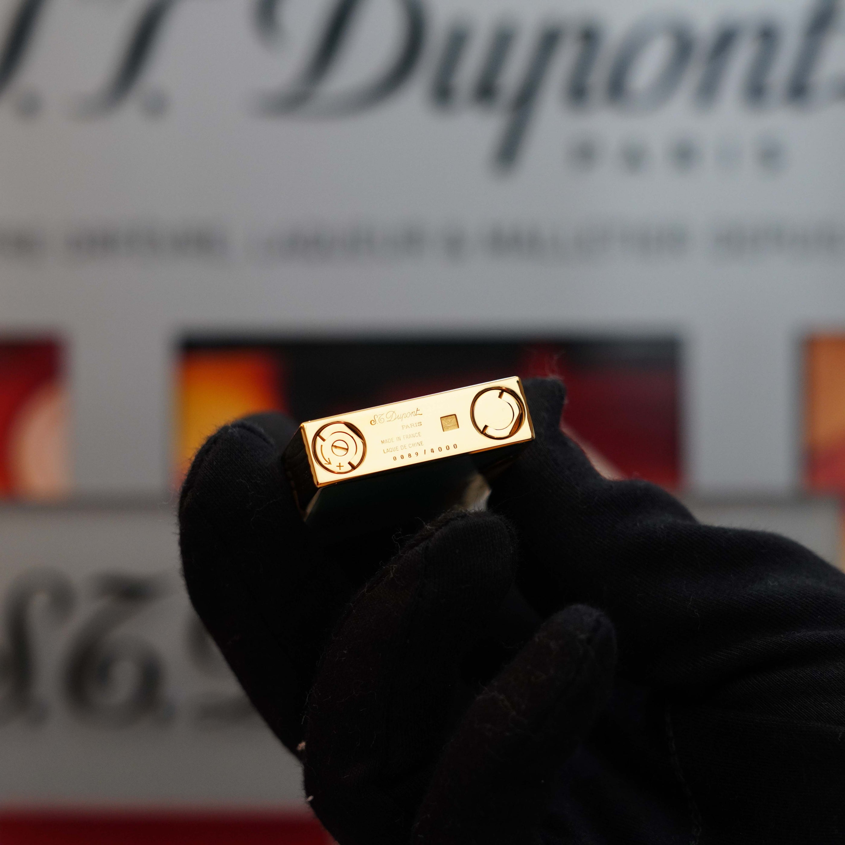 A gloved hand holds a small, rectangular gold object with intricate design details and the branding "S.T. Dupont" in the background. This exquisite piece, part of the S.T. Dupont Limited Editions, showcases a 24k gold finish reminiscent of a Vintage 1993 rare Art Nouveau early Limited Editions 24k Gold finish natural Green lacquer ligne 2 lighter's elegance.