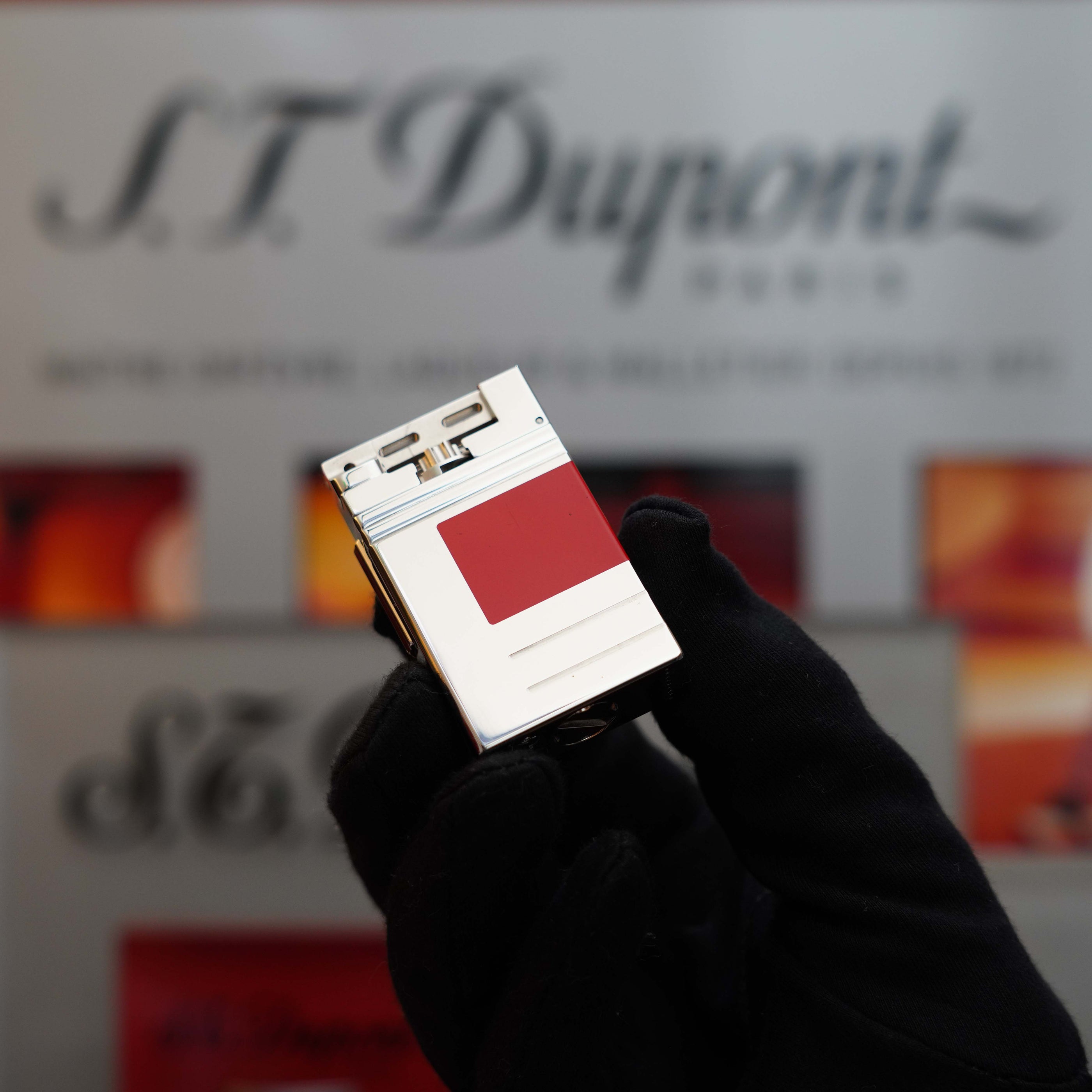 A gloved hand holding a Vintage 1999 S.T. Dupont Urban Limited Edition Platinum finish natural Red Lacquer lighter is displayed in front of a blurred S.T. Dupont sign, making for an exquisite collector's item.