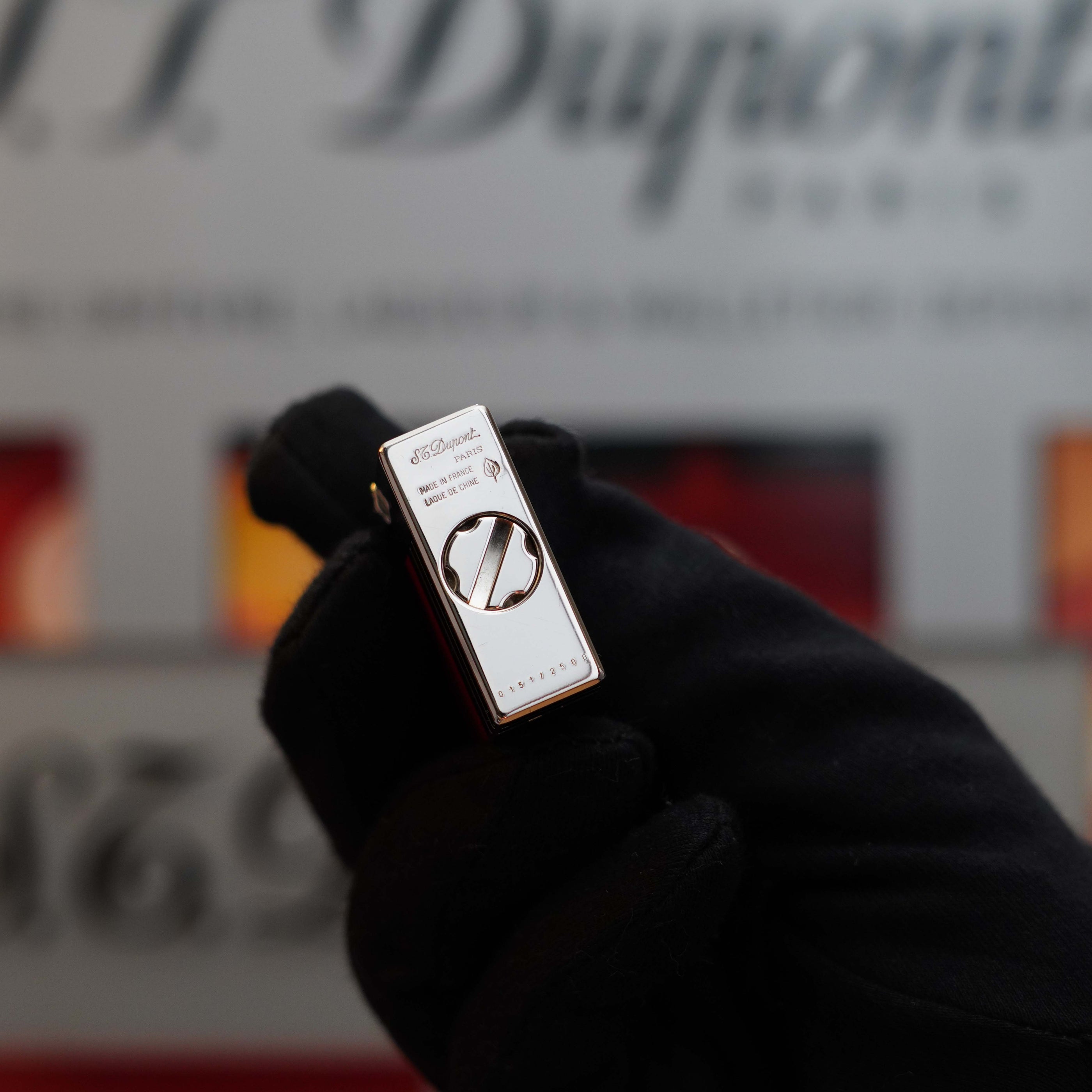 A gloved hand holds a close-up of a Vintage 1999 St Dupont Urban Limited Edition Platinum finish natural Red Lacquer lighter against a blurred background. The lighter features a sleek metallic design with the S.T. Dupont logo and some text visible, making it a perfect collector's item.