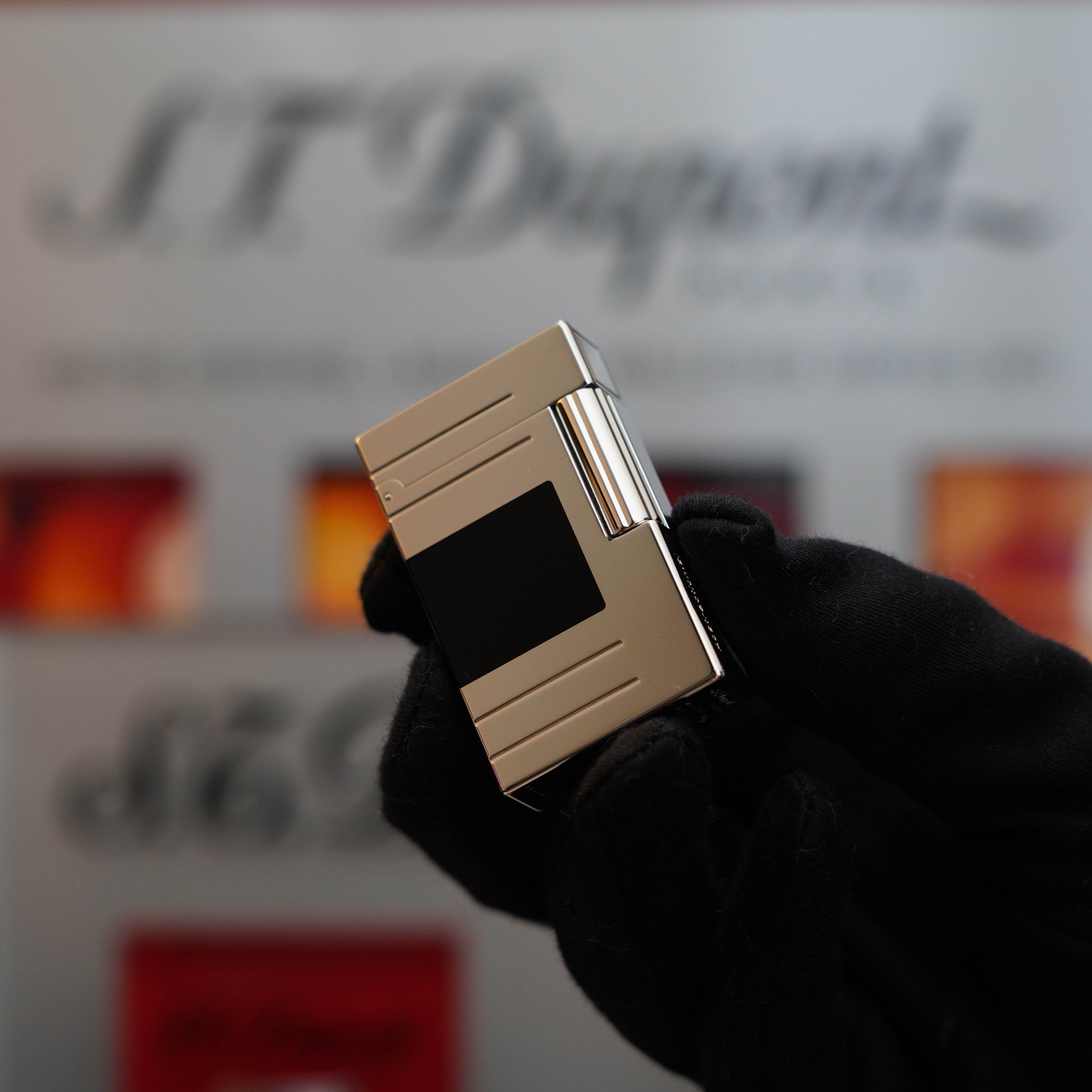 A gloved hand holds a silver rectangular lighter with a black stripe in front of a blurry backdrop featuring the logo "S.T. Dupont," showcasing the elegance of the Vintage 1999 St Dupont Ligne 1 Limited Edition Platinum finish natural Black Lacquer lighter design.