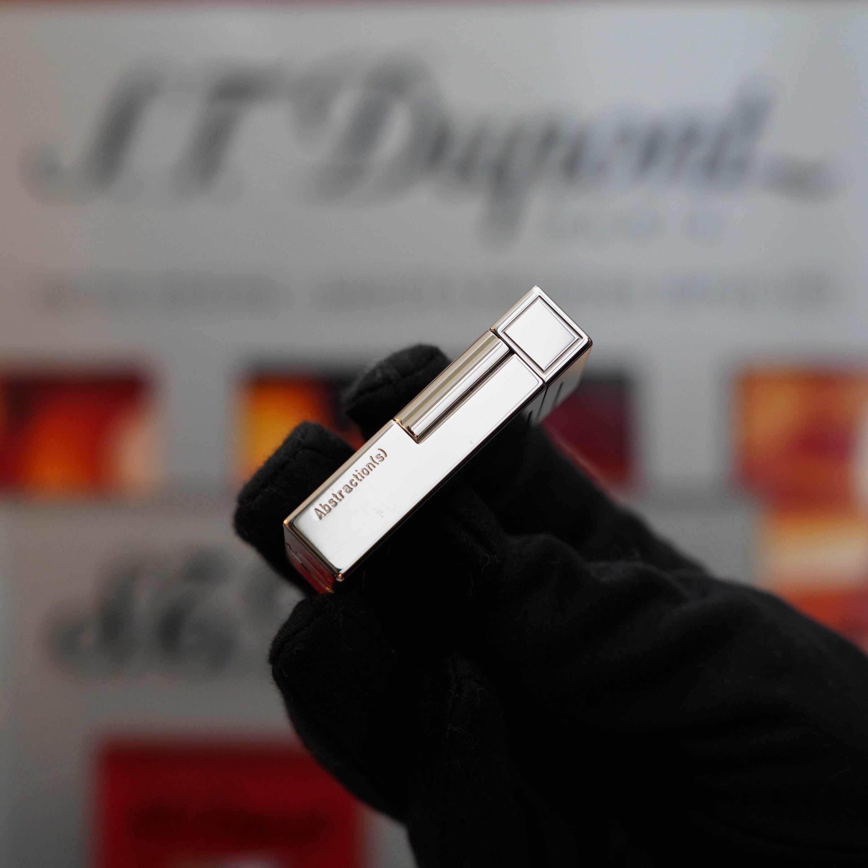 A gloved hand holds a silver cigar lighter horizontally, with "Alexander" engraved on it and a blurred "S.T. Dupont" sign in the background. The Vintage 1999 S.T. Dupont Ligne 1 Limited Edition Platinum finish natural Black Lacquer lighter exudes timeless elegance, capturing a moment of luxury and refinement.