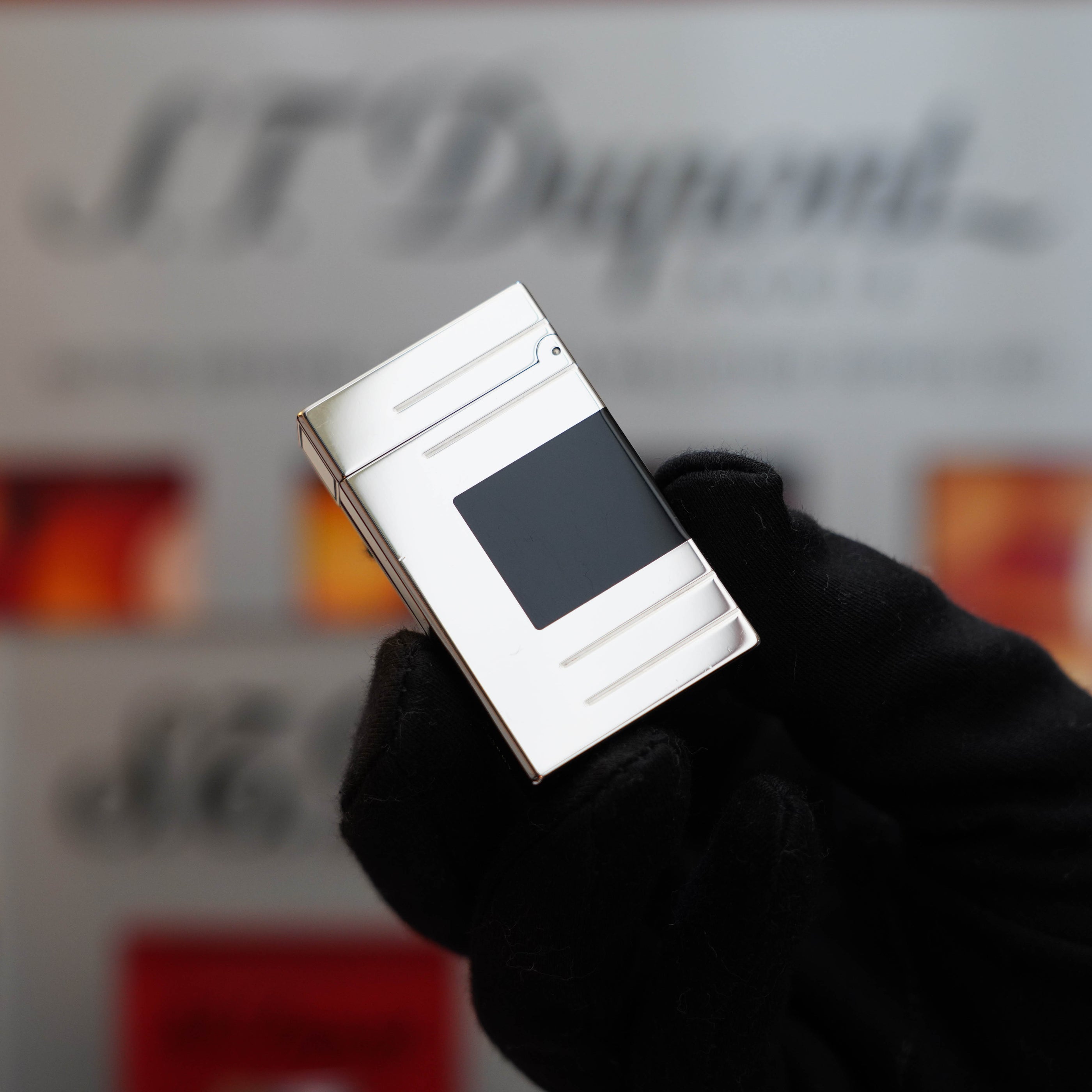 A gloved hand holds a Vintage 1999 S.T. Dupont Ligne 1 Limited Edition Platinum finish natural Black Lacquer lighter, adorned with a sleek black lacquer panel, against a blurred background featuring the brand name "S.T. Dupont.