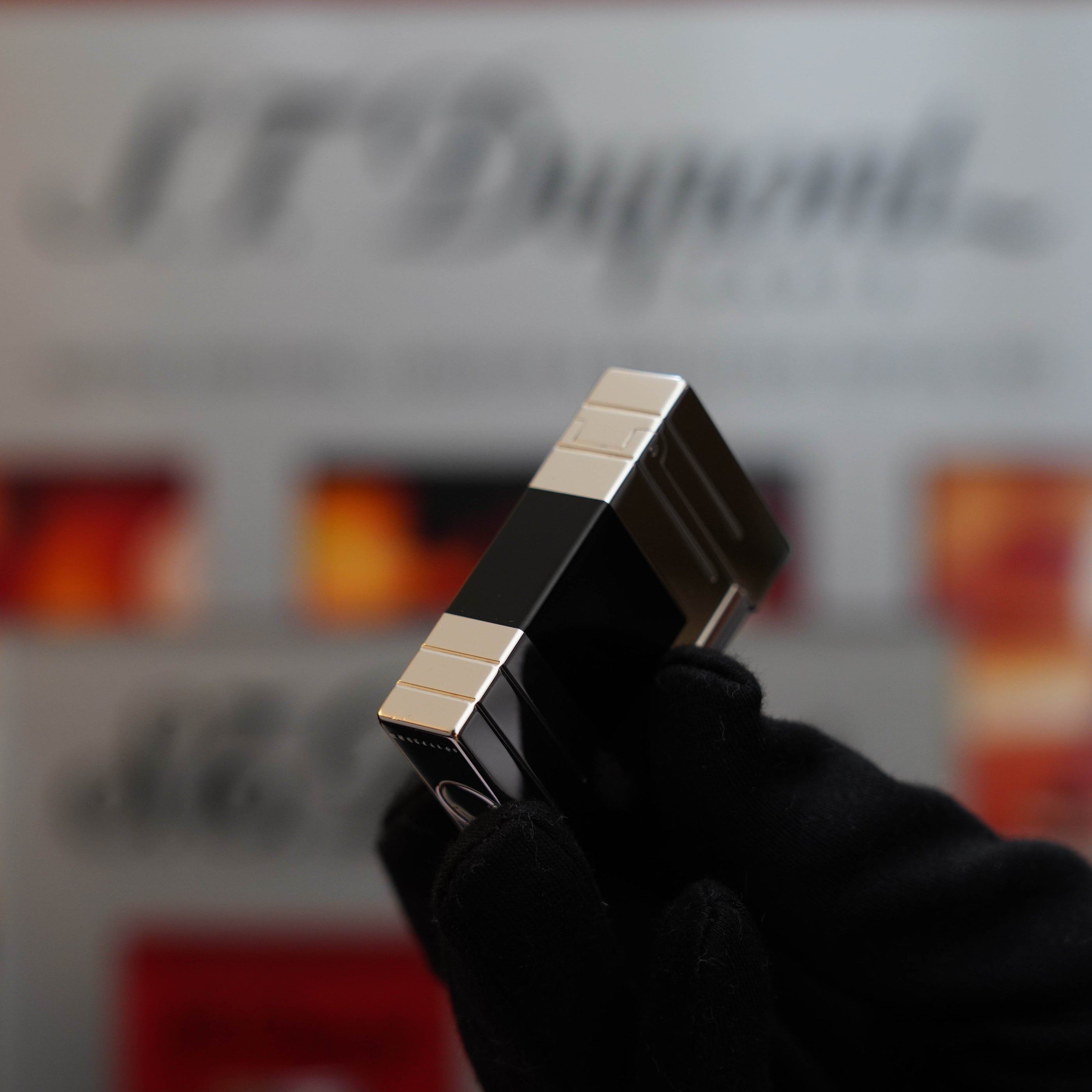 A gloved hand holds a Vintage 1999 St Dupont Ligne 1 Limited Edition Platinum finish natural Black Lacquer lighter. The background shows blurred text and images, with "S.T. Dupont" partially visible.