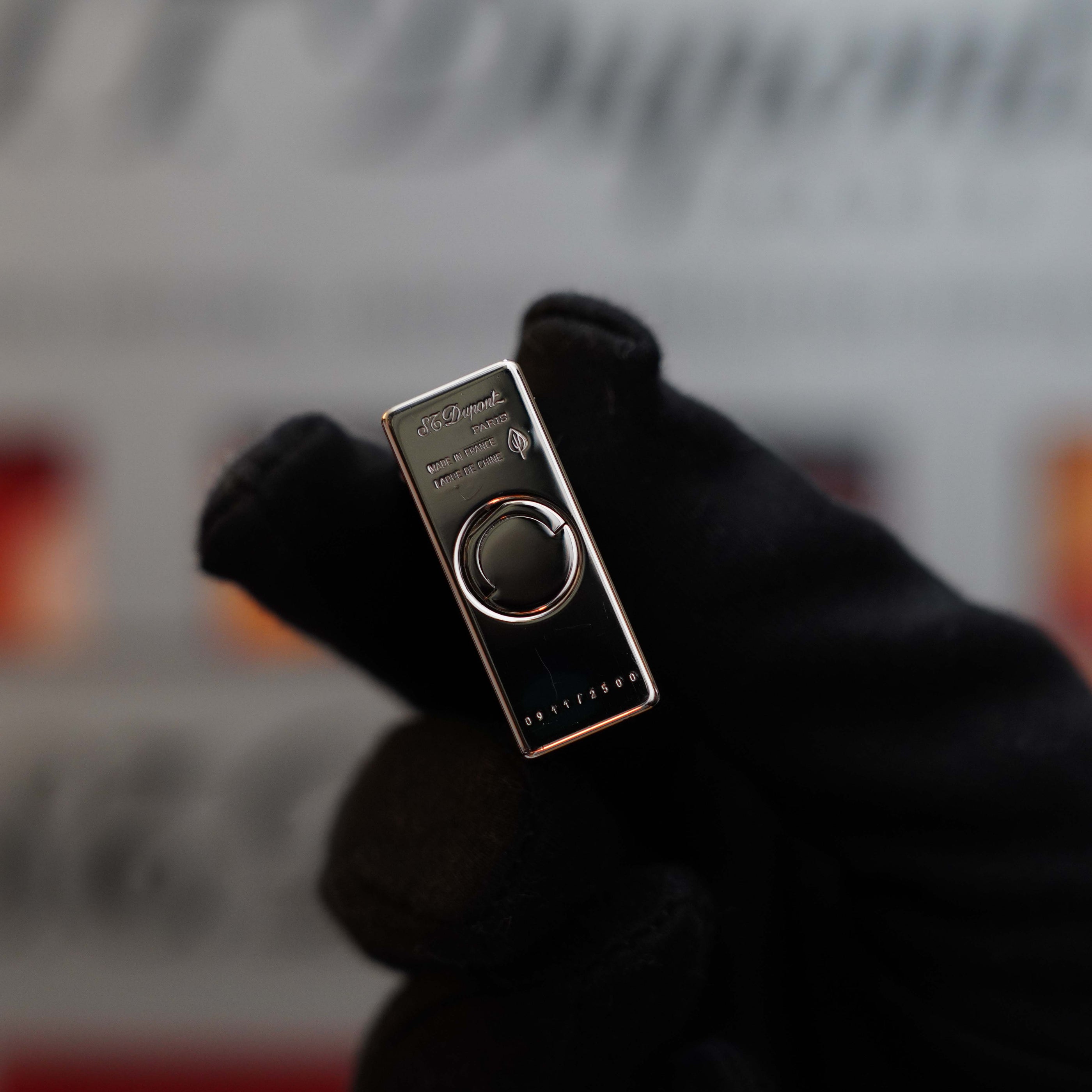 A gloved hand holds a small, rectangular Vintage 1999 S.T. Dupont Ligne 1 Limited Edition Platinum finish natural Black Lacquer lighter with a circular button in the center and embossed text. The background is blurred and features indistinct text and colors.