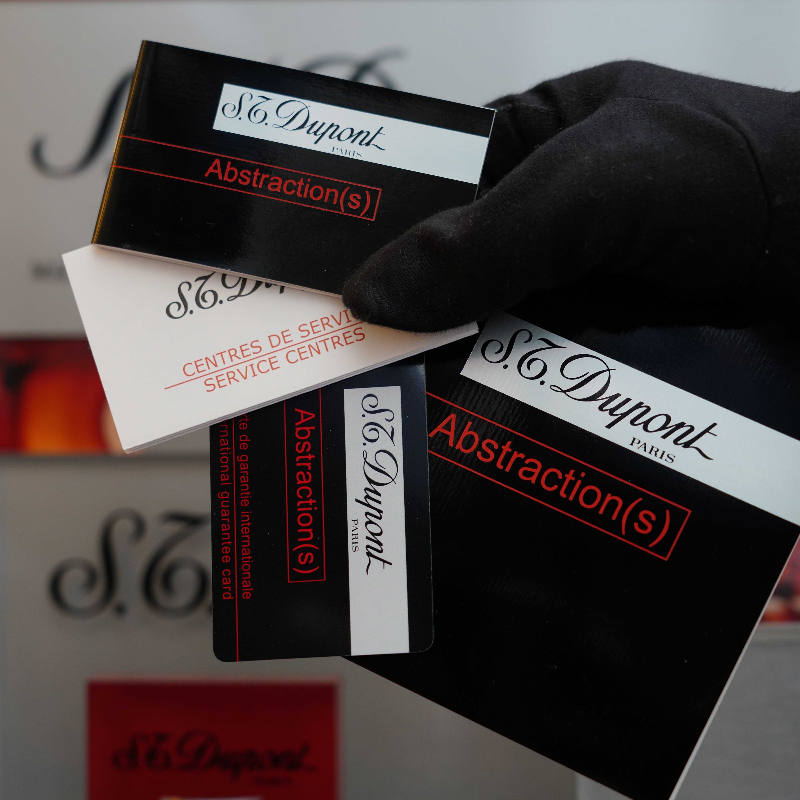 A gloved hand holds S.T. Dupont branded product packaging with the words "Abstraction(s)" and "Service Centers" visible on the boxes, hinting at a Vintage 1999 St Dupont Ligne 1 Limited Edition Platinum finish natural Black Lacquer lighter. Logos and text are seen in the background.