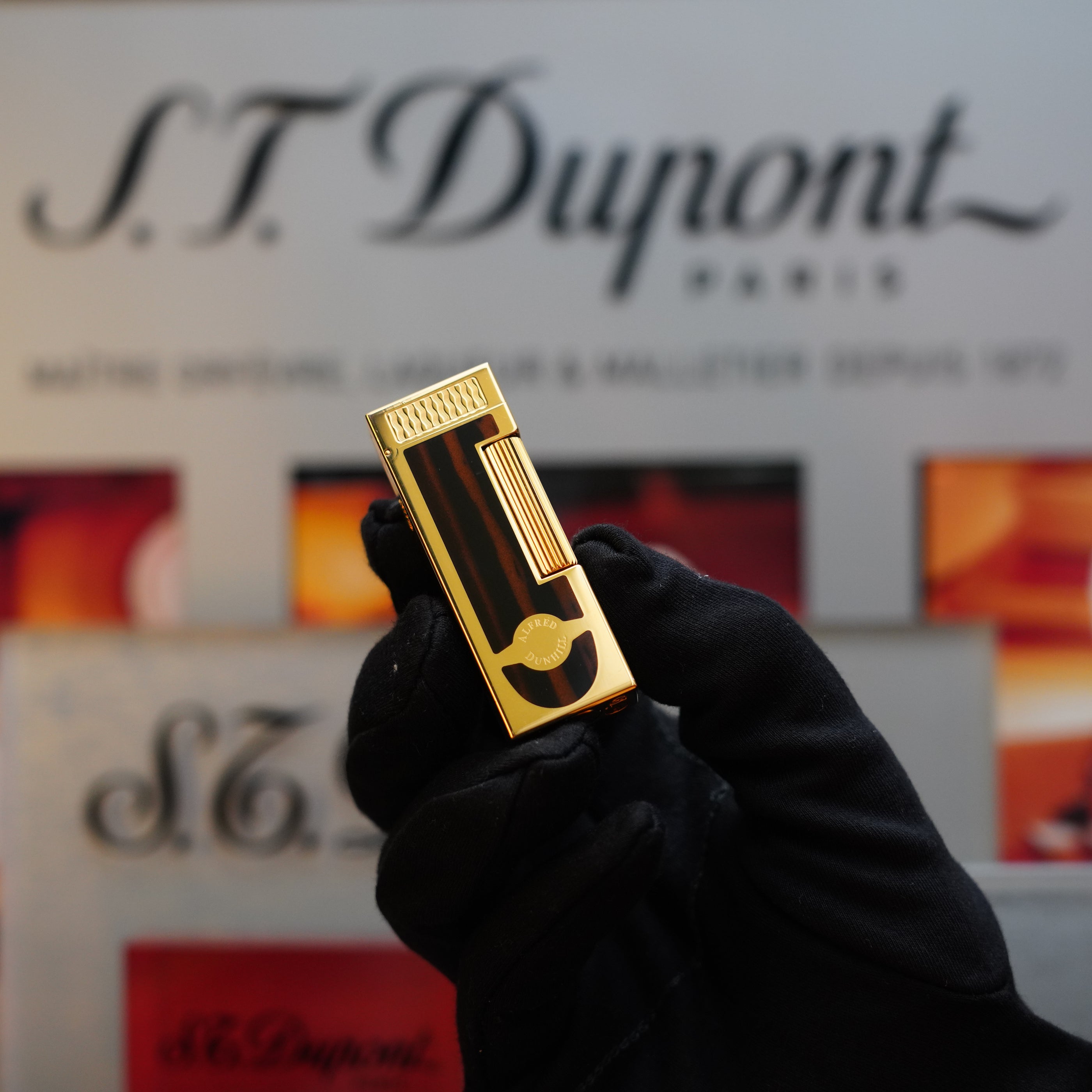 Close-up of a gloved hand holding a shiny gold lighter with a 24k gold finish, in front of a Dunhill branded background. The 1990 Dunhill Cigar Rollagas Macassar is a rare collector's item that captivates elegance and luxury at its finest.