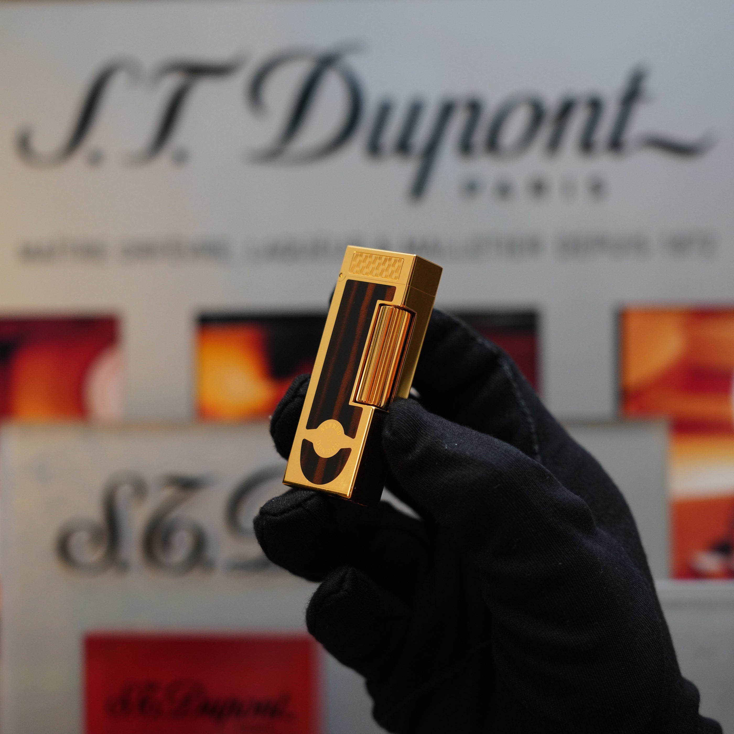 A gloved hand holding a rare collector's item, the gold-finished Dunhill 1990 Cigar Rollagas Macassar lighter, against a blurred Dunhill branded background.