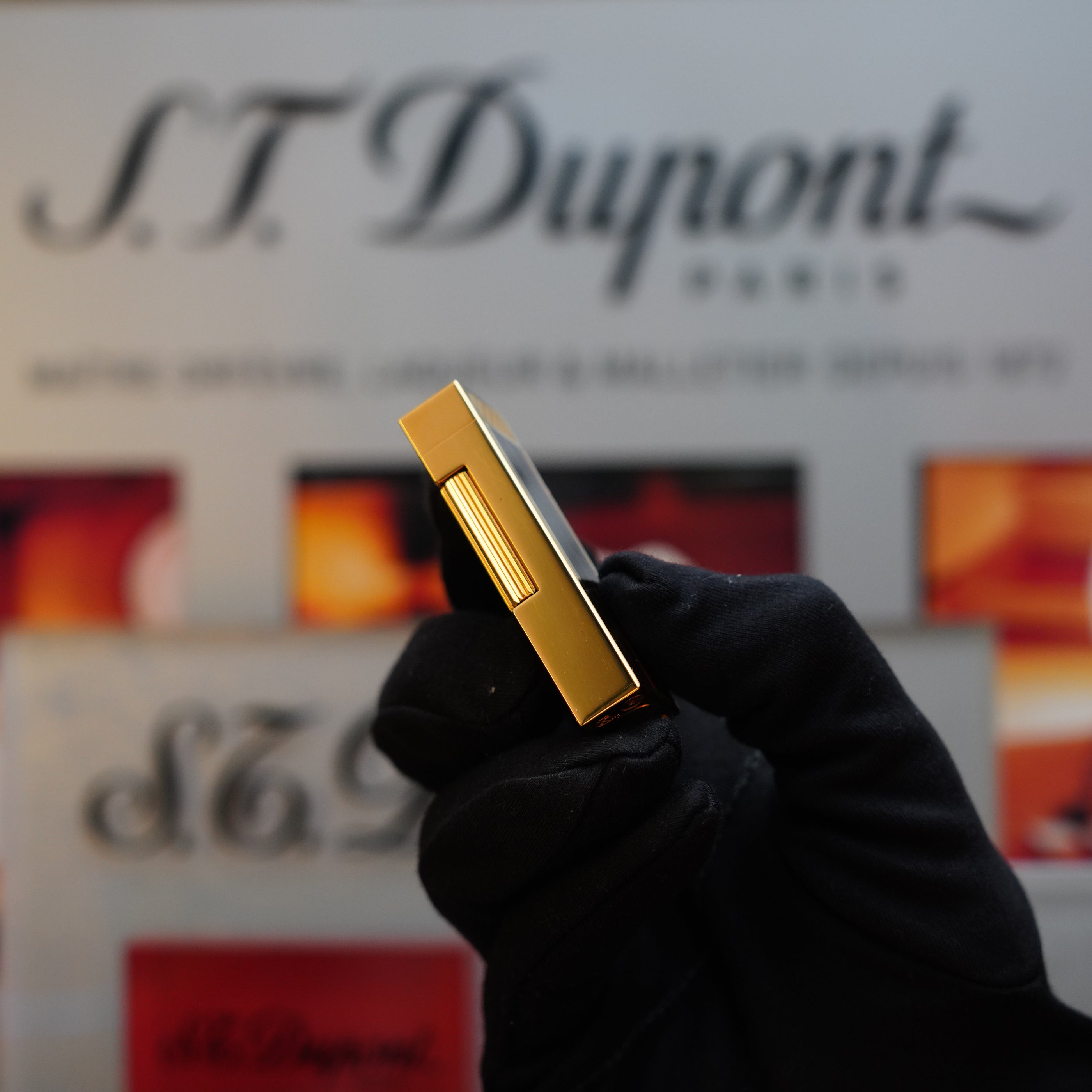 A gloved hand holds a gold-colored 1990 Dunhill Cigar Rollagas Macassar 24k Gold Finish lighter, with the Dunhill logo visible in the blurred background, reminiscent of a rare collector's item.