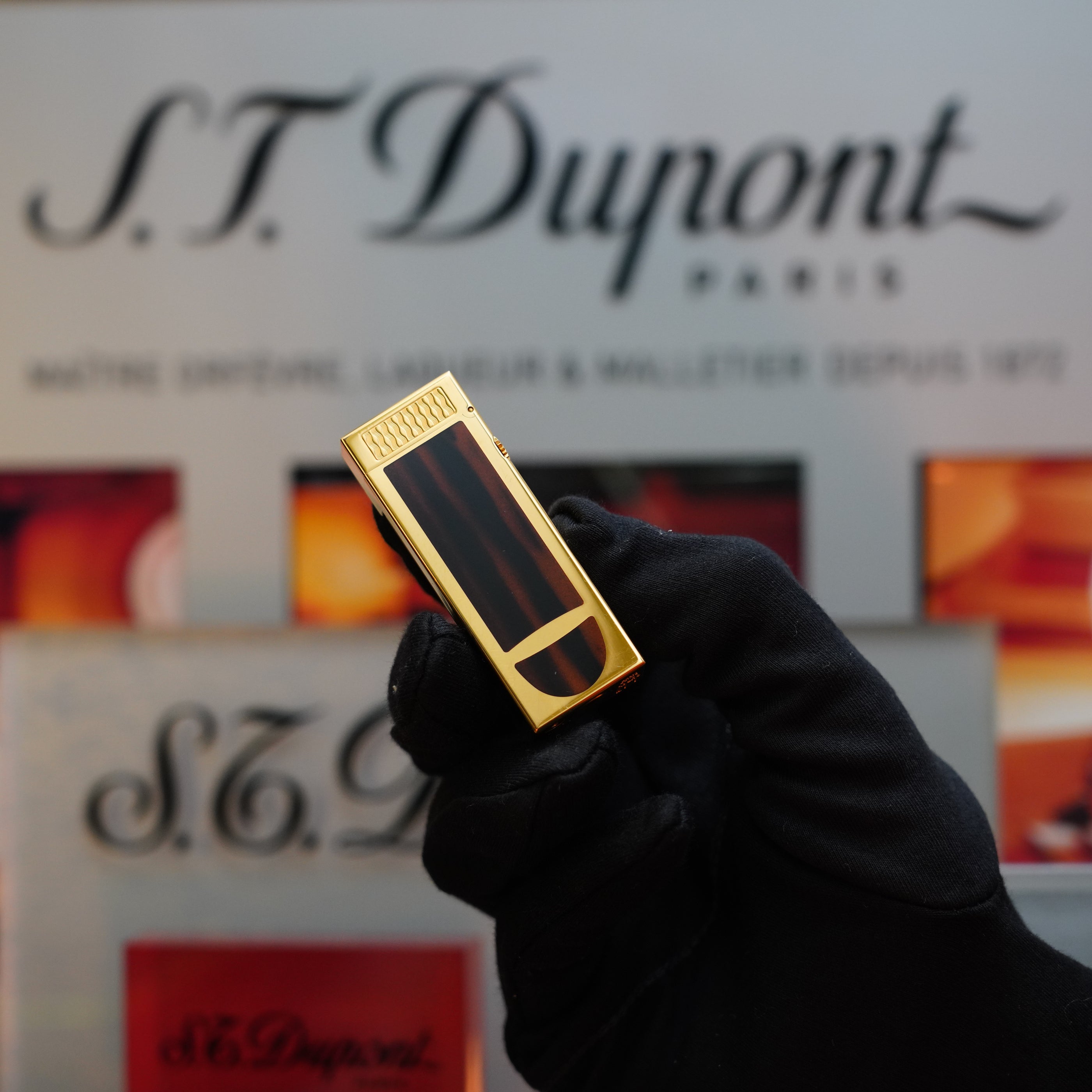 A gloved hand holds a rectangular, 1990 Dunhill Cigar Rollagas Macassar 24k Gold Finish lighter in front of a blurred background featuring the brand name "Dunhill.