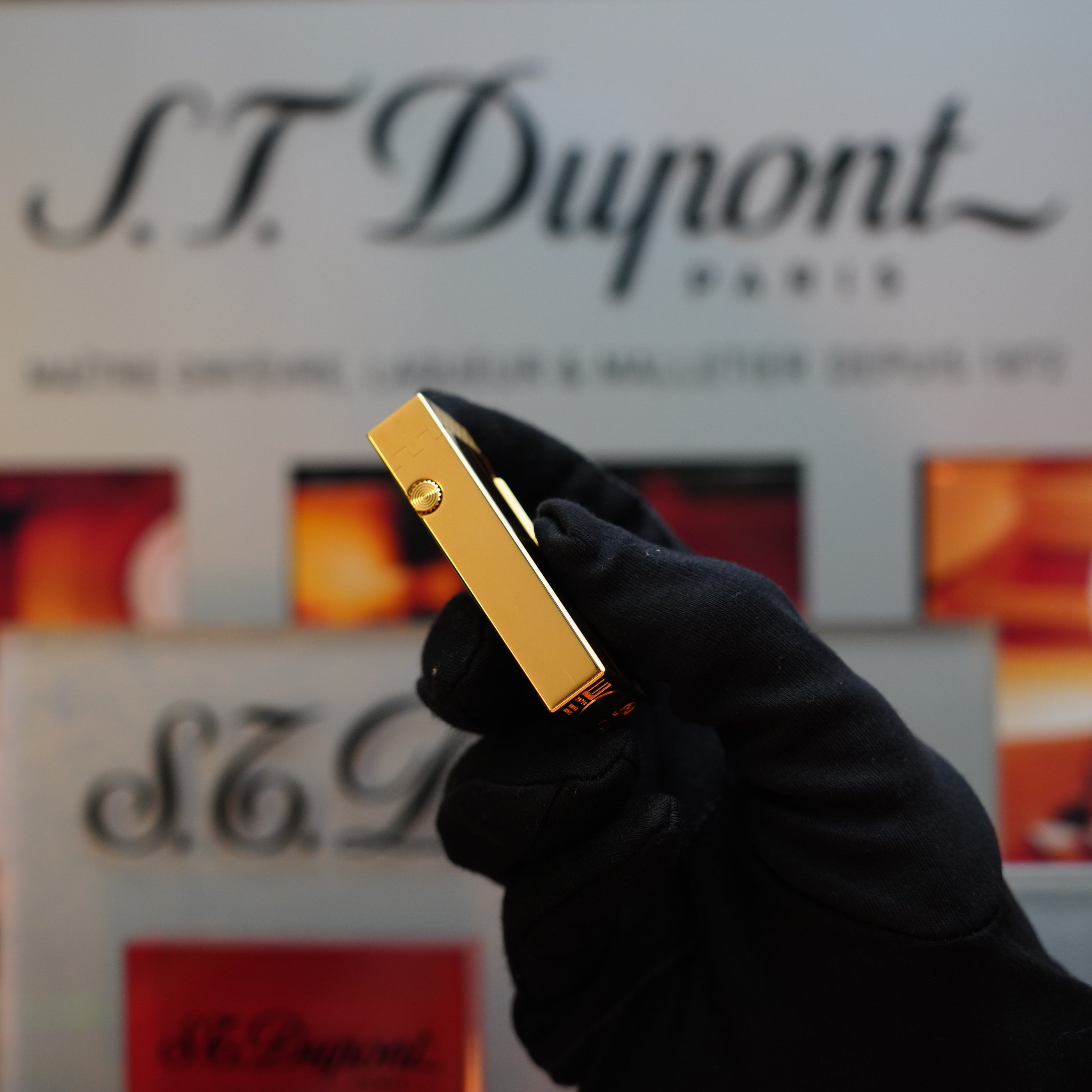 A gloved hand holds a gold-colored Dunhill item, reminiscent of a vintage S.T. Dupont lighter. The background features a blurred Dunhill logo and some colorful designs, highlighting this 1990 Dunhill Cigar Rollagas Macassar 24k Gold Finish lighter as a rare collector's item.