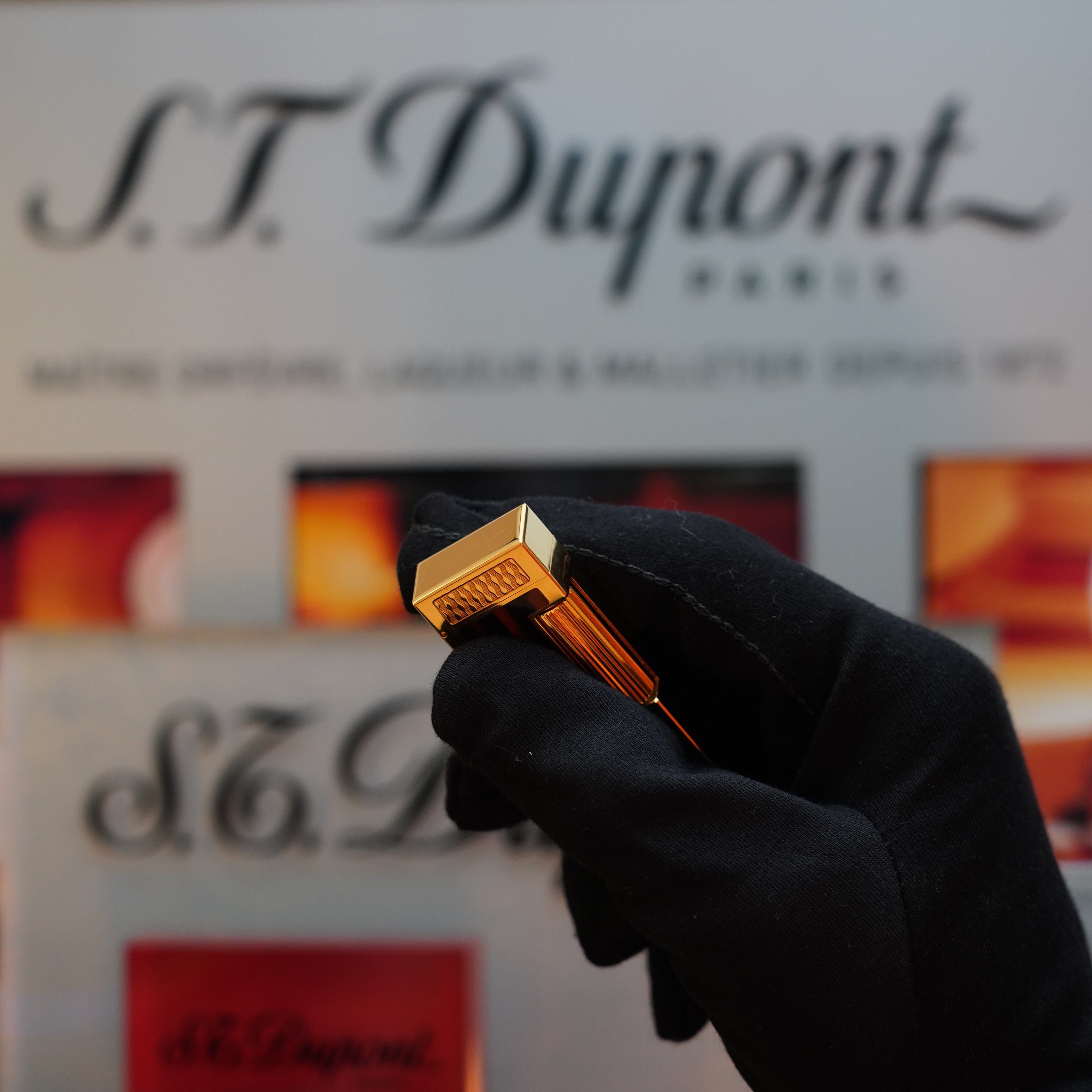 A gloved hand holds a 1990 Dunhill Cigar Rollagas Macassar 24k Gold Finish lighter, an exquisite Dunhill piece, in front of a display sign with the Dunhill logo.