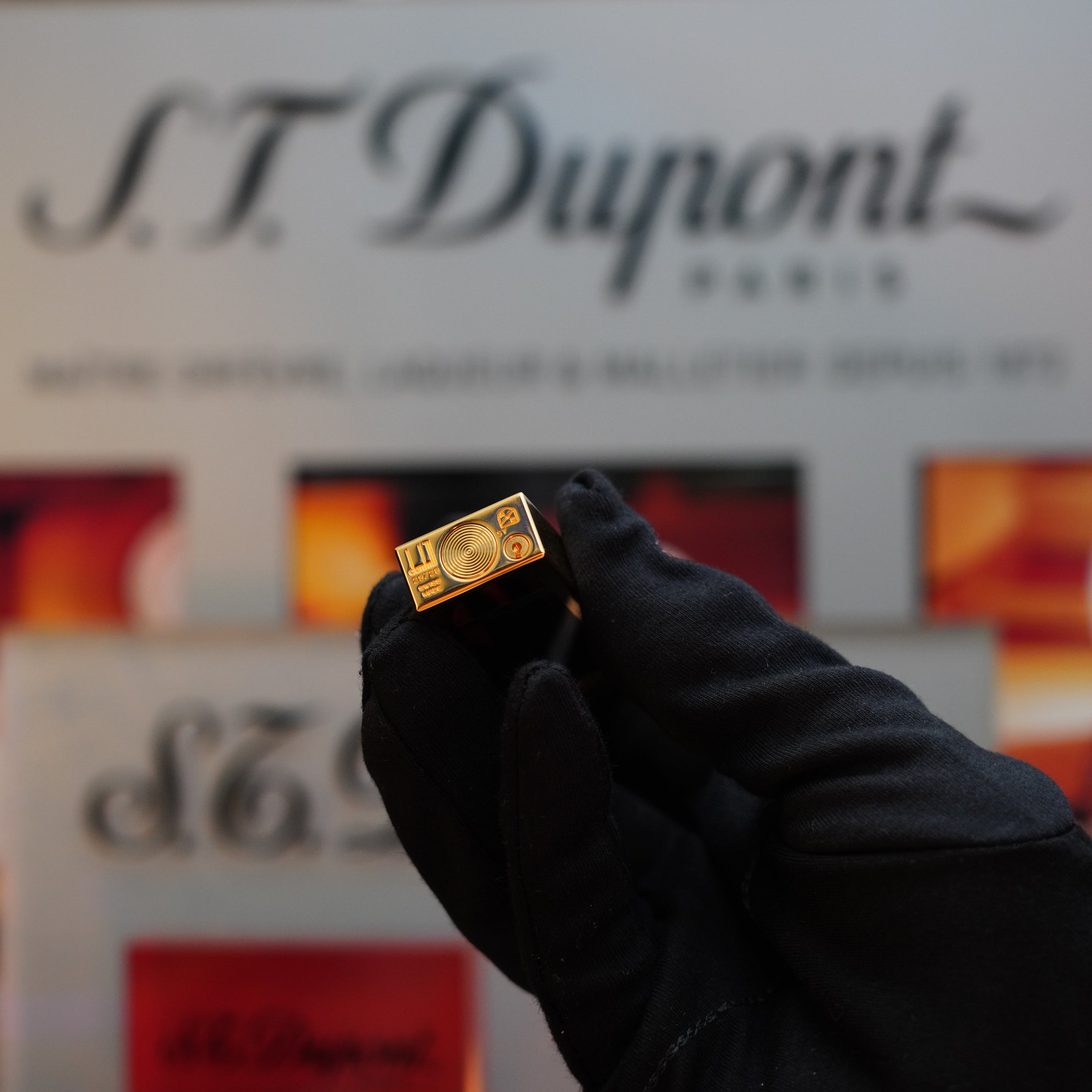 A gloved hand holds a rare collector's item—a 1990 Dunhill Cigar Rollagas Macassar with a 24k gold finish—in front of a blurred background featuring the Dunhill logo and various lighter models.