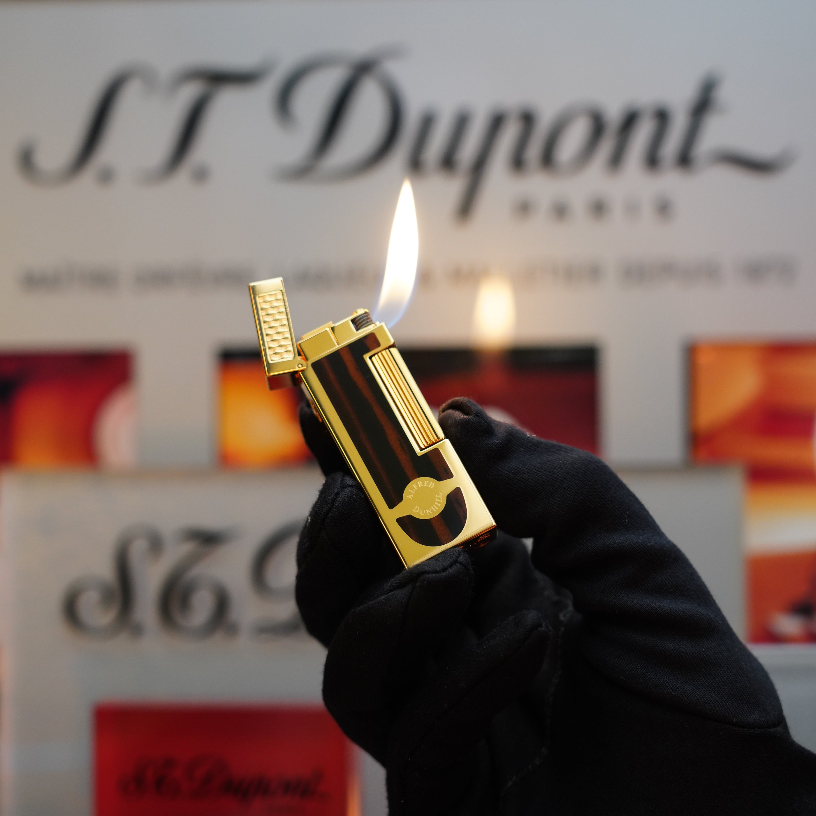 A gloved hand holds a lit 1990 Dunhill Cigar Rollagas Macassar 24k Gold Finish lighter with a gold and black design, reminiscent of a Rare Collector's Item. The Dunhill brand's logo and signage are visible in the background.