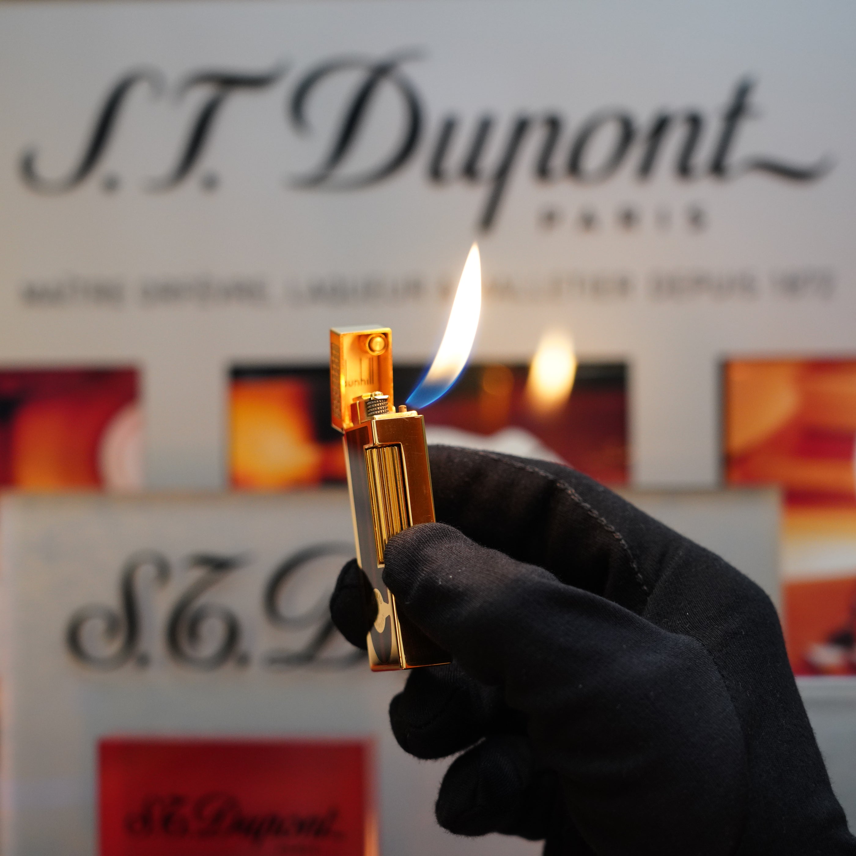 A gloved hand holds a lit 1990 Dunhill Cigar Rollagas Macassar 24k Gold Finish lighter in front of a sign reading "S.T. Dupont Paris." The flame is visible against the blurred background, highlighting this rare collector's item.