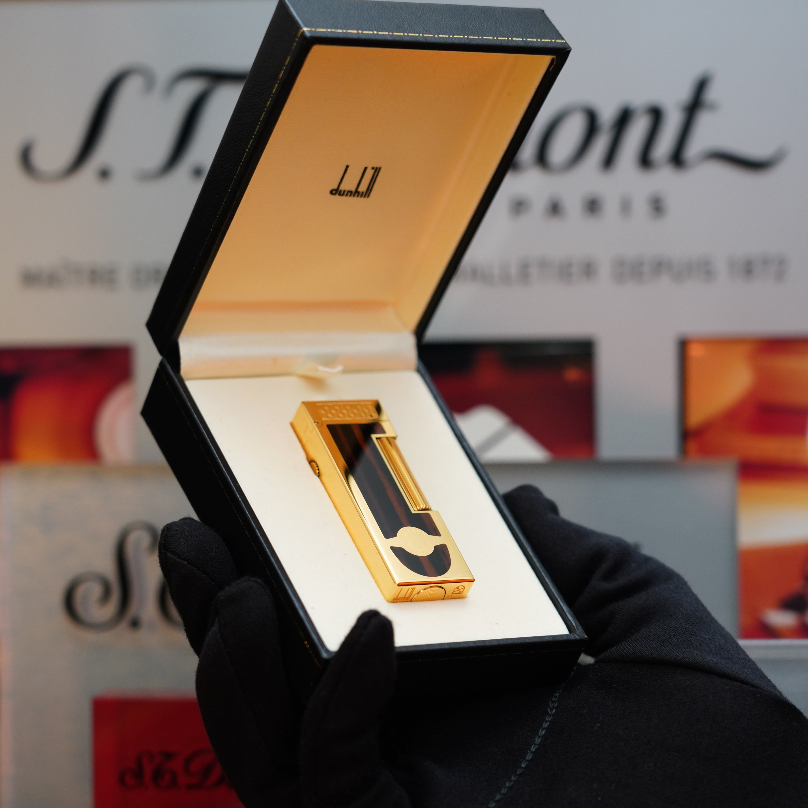 A gloved hand holds an open black and gold box containing a luxury S.T. Dupont lighter, set against a blurred background featuring S.T. Dupont branding, evoking the elegance of the 1990 Dunhill Cigar Rollagas Macassar 24k Gold Finish by Dunhill.