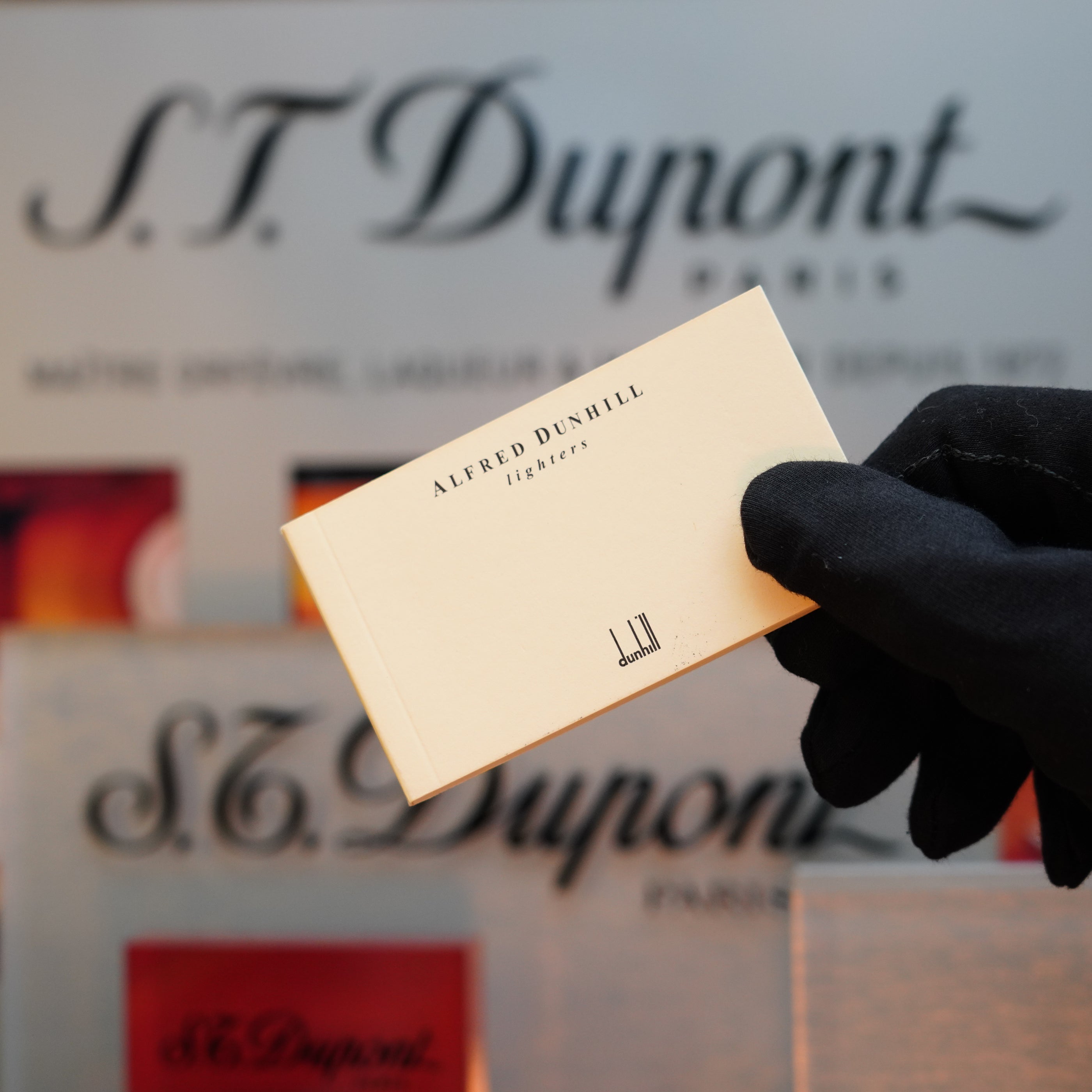 A gloved hand displays a card from Alfred Dunhill lighters against a backdrop featuring an S.T. Dupont Paris sign, drawing attention to a rare collector's item— the exquisite 1990 Dunhill Cigar Rollagas Macassar with a 24k gold finish.