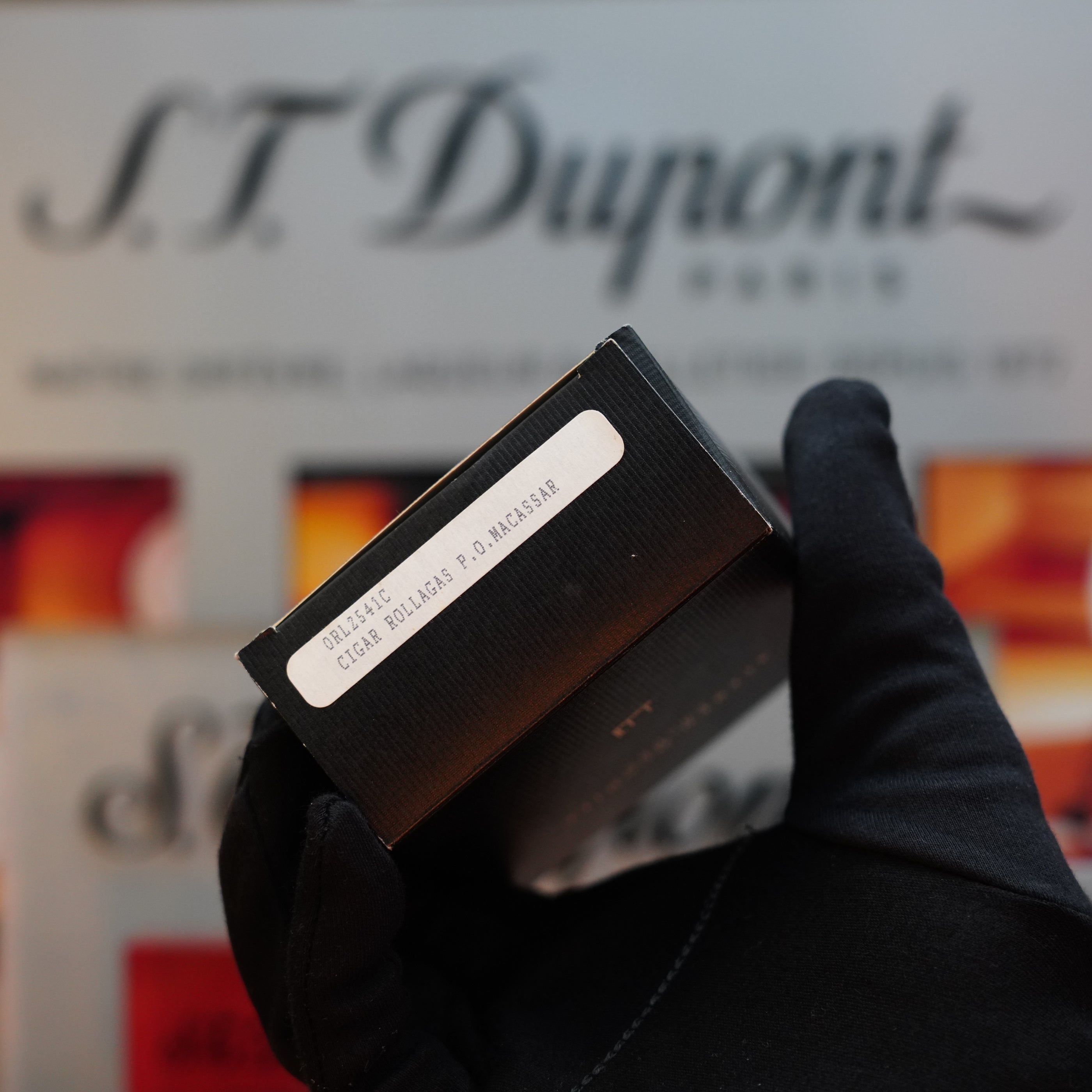 A person wearing a black glove holds a small black box with a label reading "1990 Dunhill Cigar Rollagas Macassar 24k Gold Finish." The background subtly features a blurred "Dunhill" logo, evoking the allure of a Rare Collector's Item.