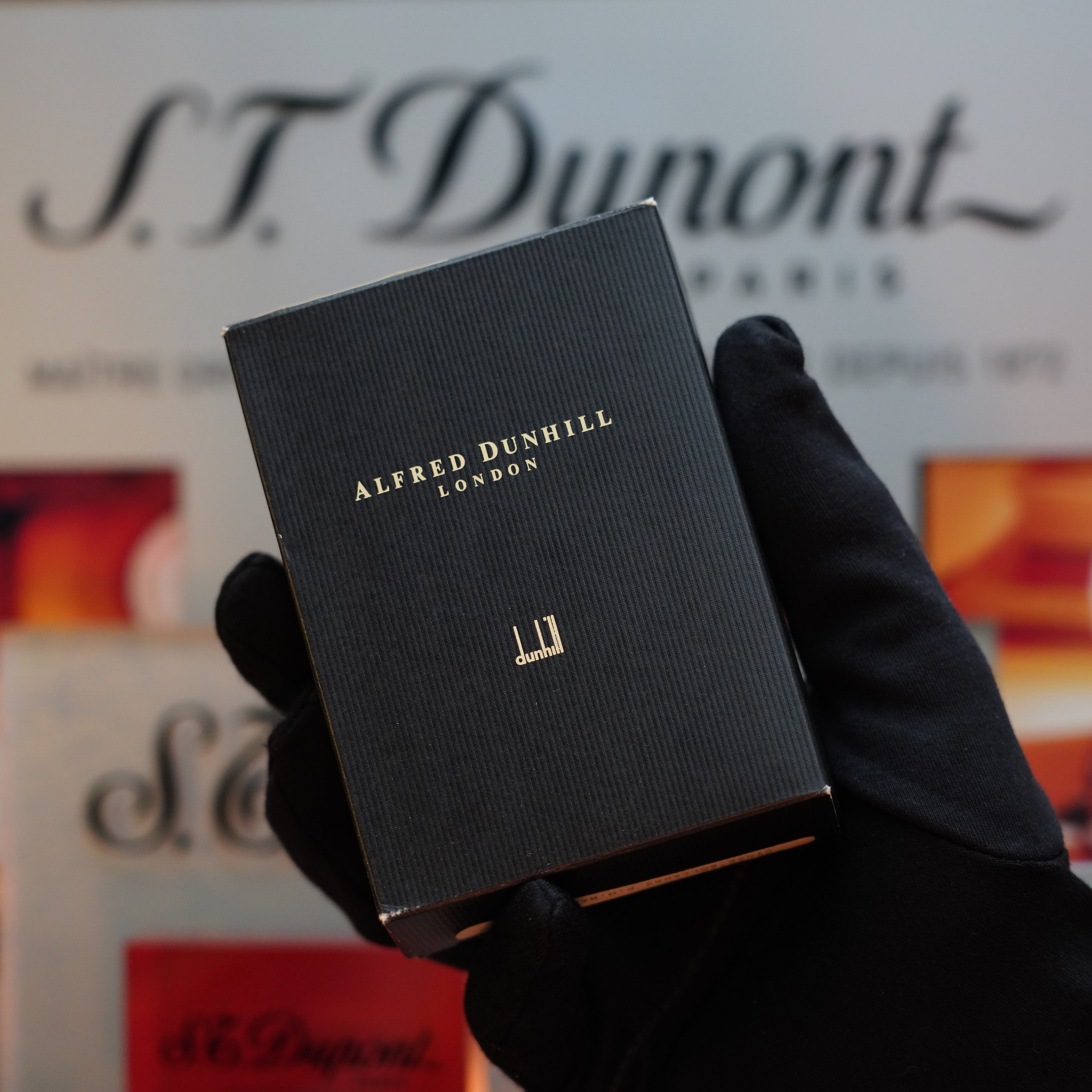 A gloved hand holds a black box adorned with gold lettering that reads "Dunhill" in front of a blurred backdrop with the text "S.T. Dupont Paris." This rare collector's item, the 1990 Dunhill Cigar Rollagas Macassar 24k Gold Finish, exudes timeless elegance.